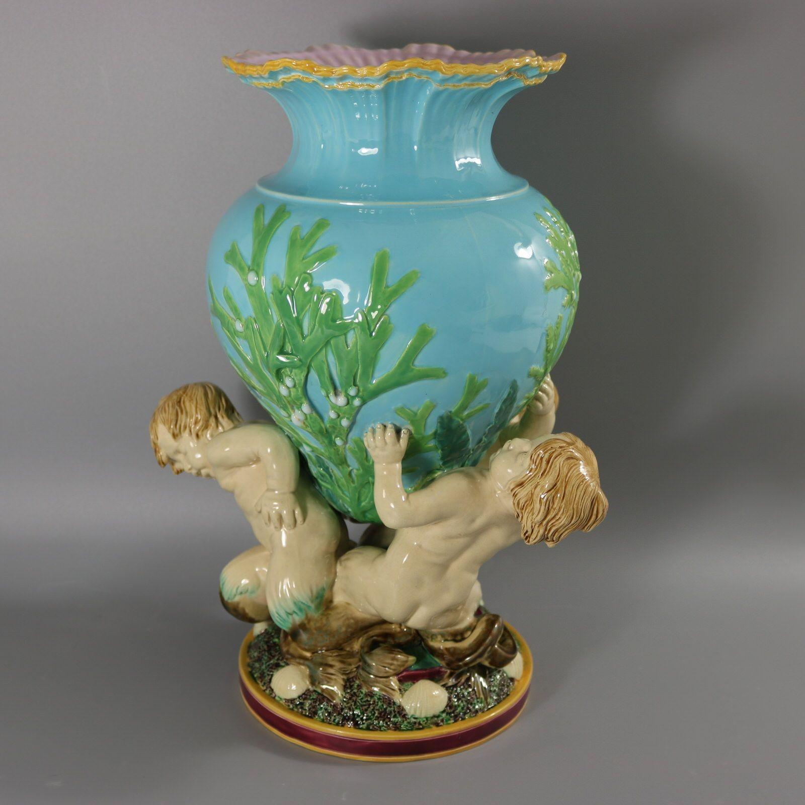 Vase en majolique de Minton qui représente trois merboys soutenant un récipient orné d'algues. Le bord du vase est modelé pour représenter des vagues déferlantes. Version avec fond turquoise. Coloration : turquoise, vert, crème, sont prédominants.