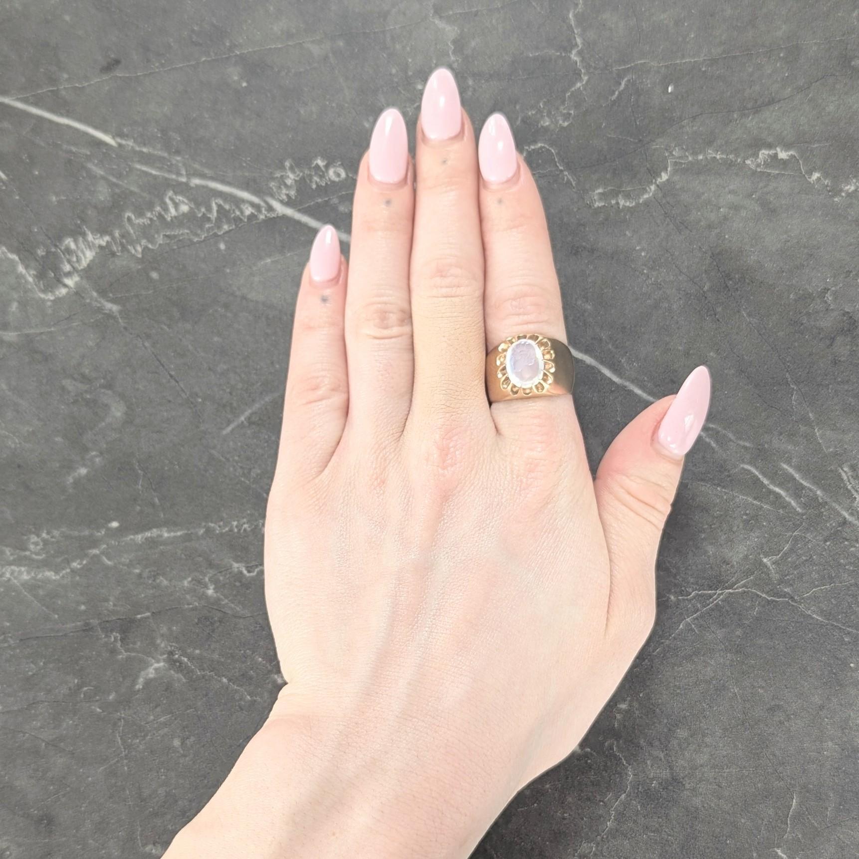 Centré sur une pierre de lune de forme ovale mesurant 8,5 x 12,0 mm
Incolore avec adularescence bleue vive et sculpté en creux.
Représentation d'une femme de profil de style grec avec bandeau
Serti de griffes de style belcher profondément