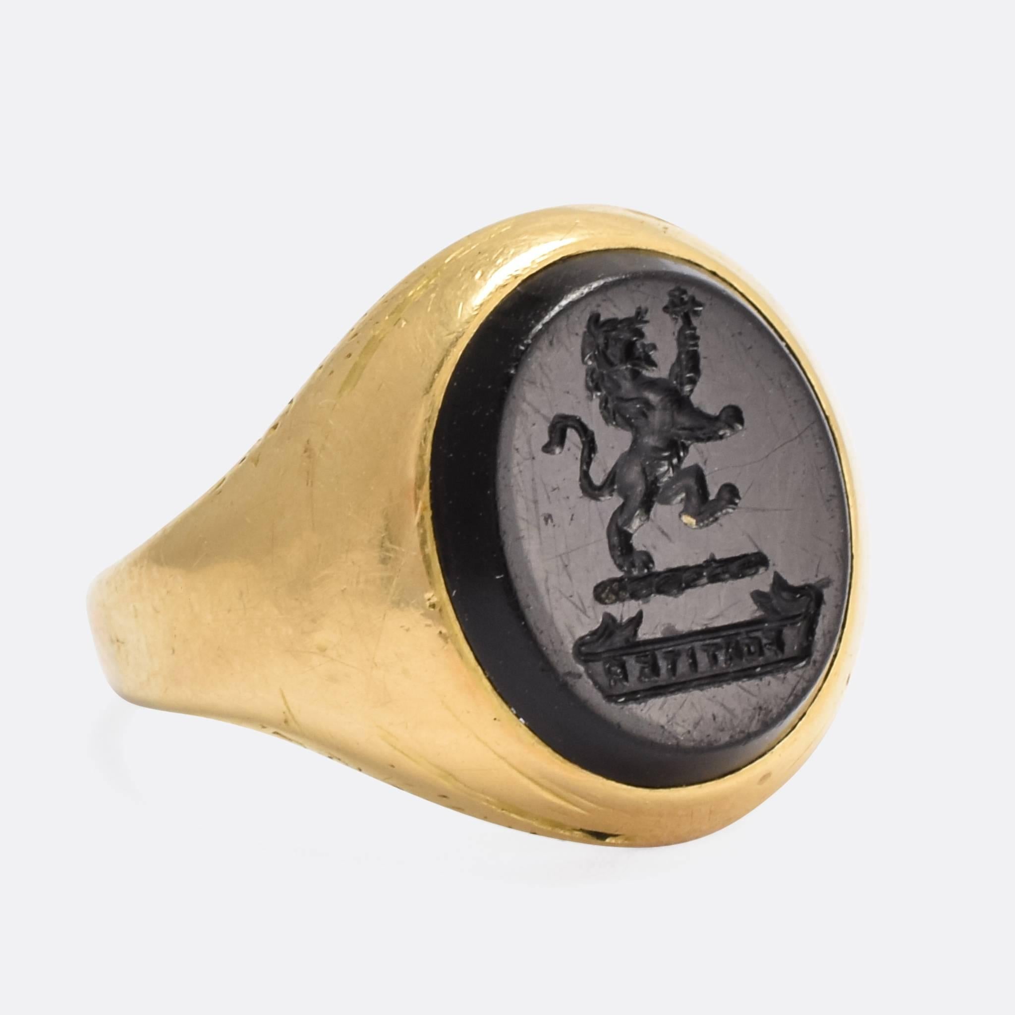 A cool antique signet ring set with an oval onyx panel, carved with a heraldic intaglio crest. Depicted is a Lion Rampant, holding a cross in its hand, above the Latin motto: 
