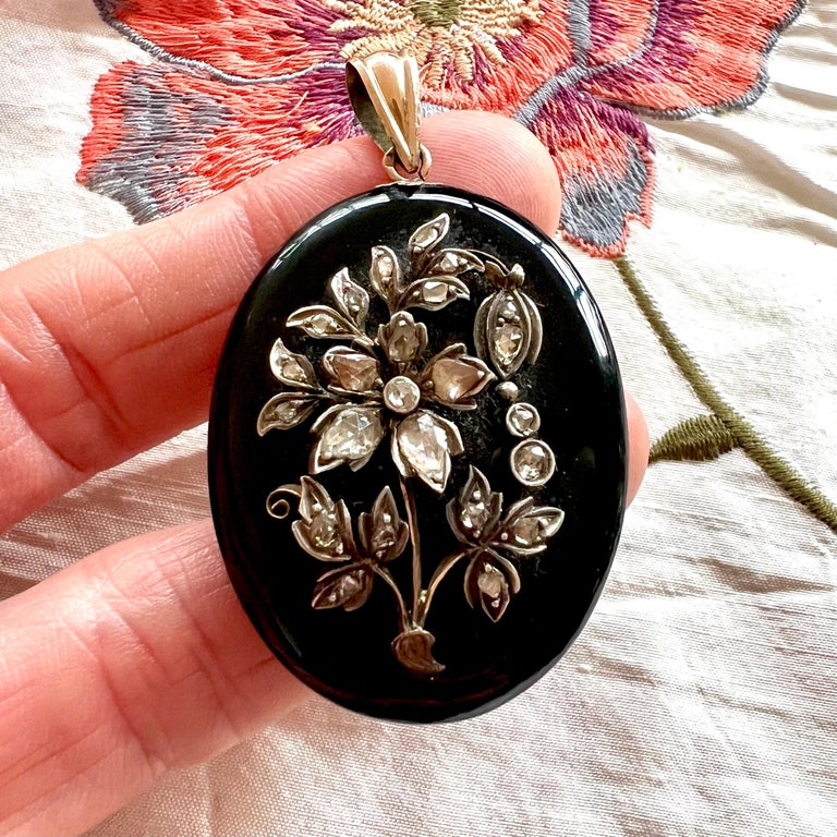 A beautiful Late Victorian onyx and diamonds mourning locket pendant. On top, the locket pendant is embellished with silver flowers and leaves which are set with rose cut diamonds. The onyx is oval-shaped and is being held by a 14 karat gold bail. A