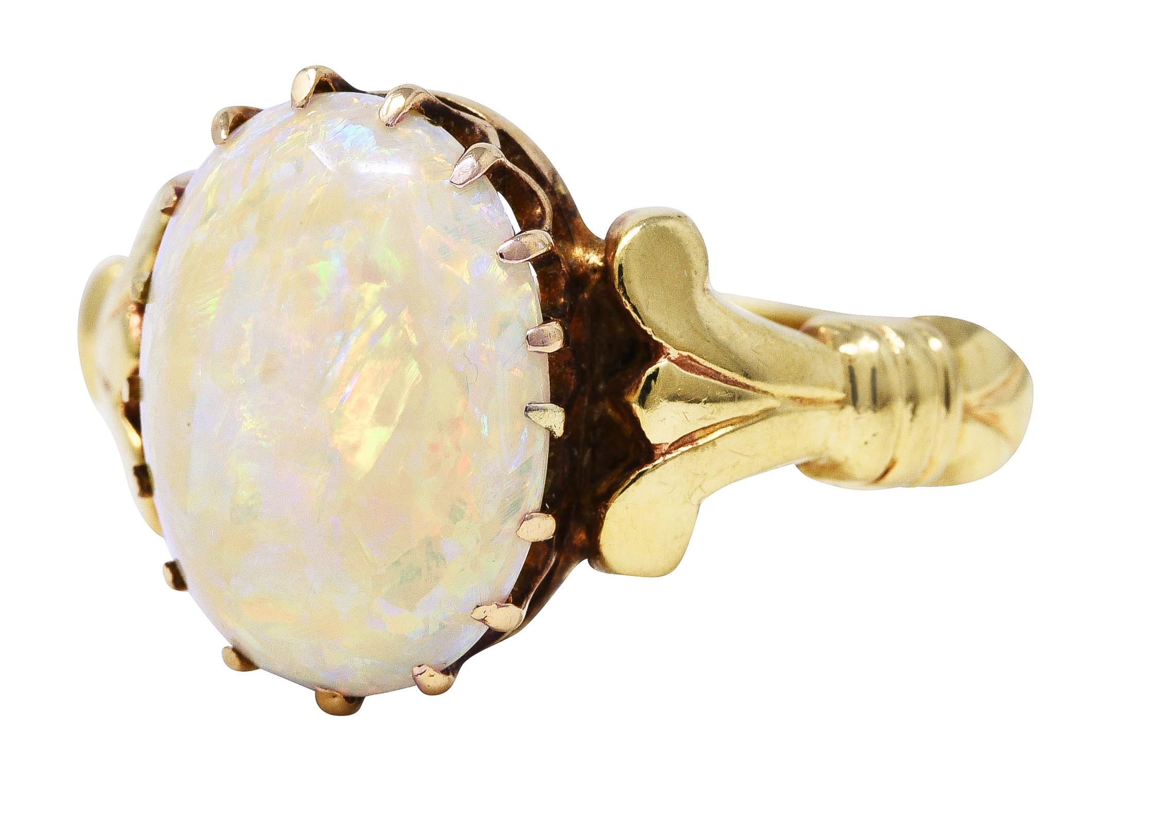 Ring centers a 12.0 x 9.0 mm oval opal cabochon - translucent white in body color with spectral play-of-color. Prong set in a scalloped gallery and flanked by grooved lotus motif shoulders with fluted accents. Tested as 14 karat gold. Circa: 1880's.