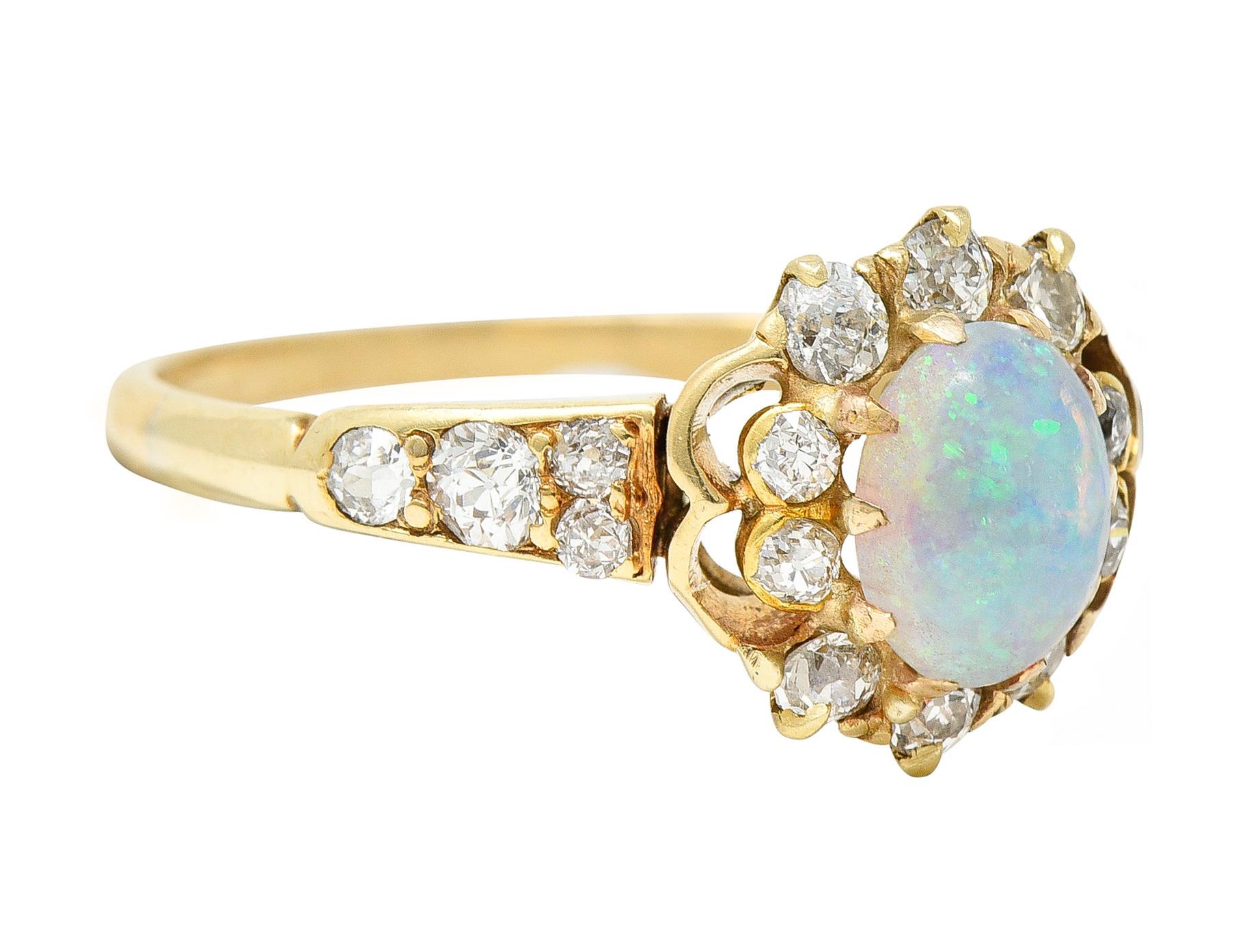 Belcher style cluster ring centering an oval opal cabochon measuring approximately 7.0 x 5.0 mm. Translucent white in body color with strong green, violet, and blue play-of color and a moderately strong orange glow. Surrounded by a halo of old