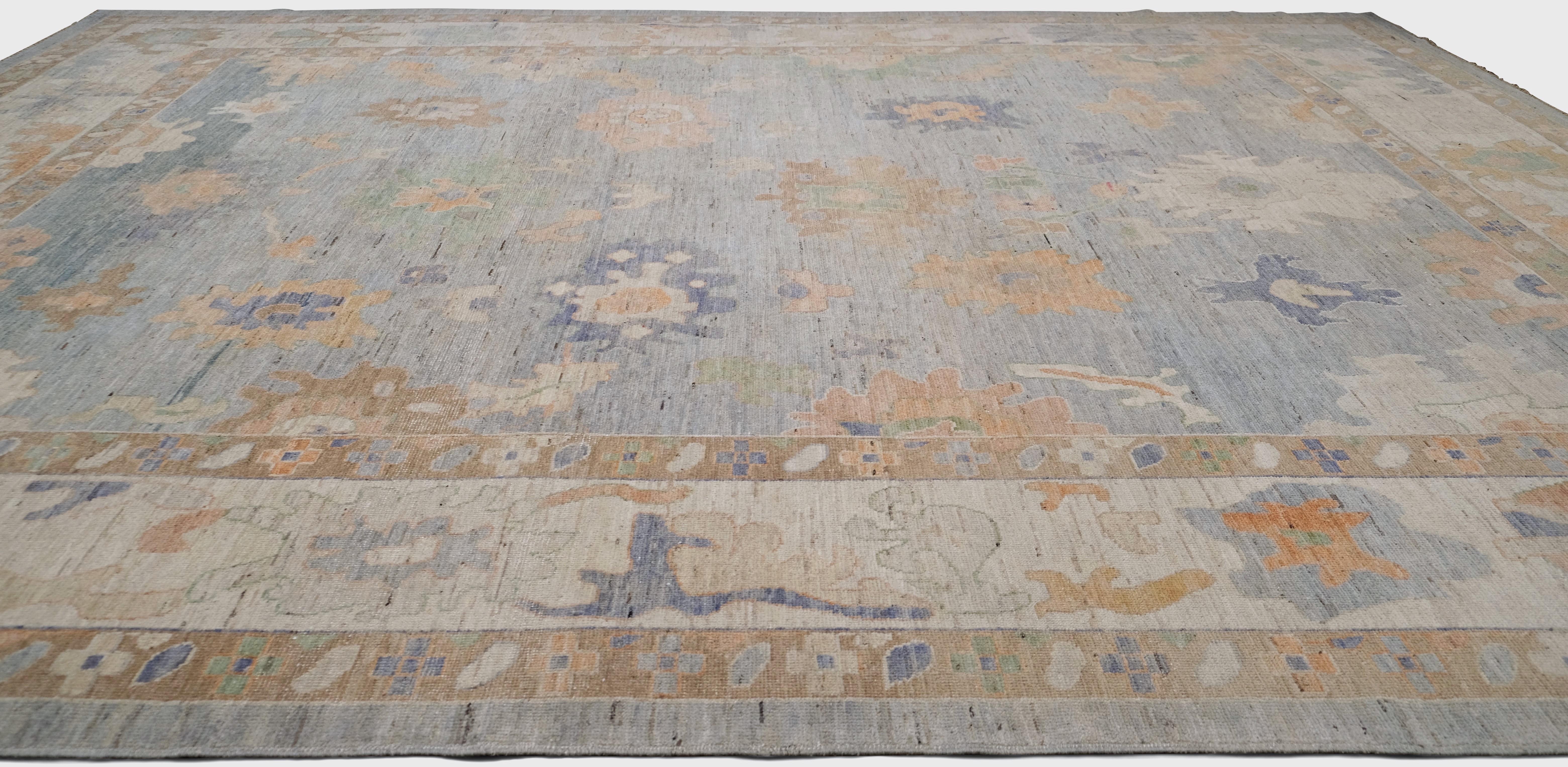 Dieser handgeknüpfte Wollteppich ist ein wunderschönes Stück im Oushak-Stil mit einem eleganten floralen Muster und einer sanften Pastellfarbpalette. Das hellblaue Feld, das mit Elfenbein und Beige akzentuiert ist, verleiht ihm einen heiteren und