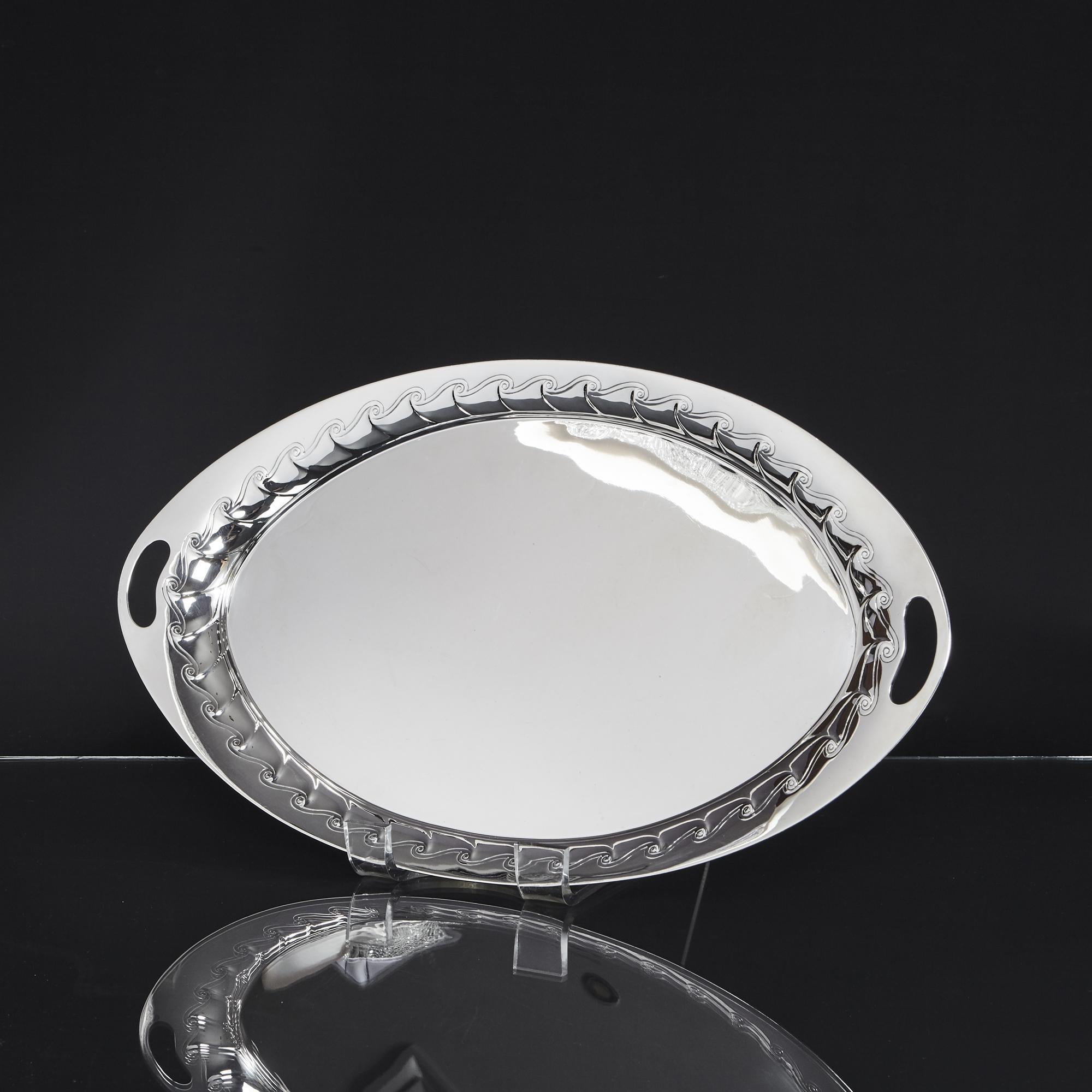 Graceful oval tray in sterling silver with a flared rim that is embossed and engraved with slanted fluting and wave-like scrolls. It has two handles incorporated into the rim.
This tray would be ideal for serving drinks or to take a three piece tea