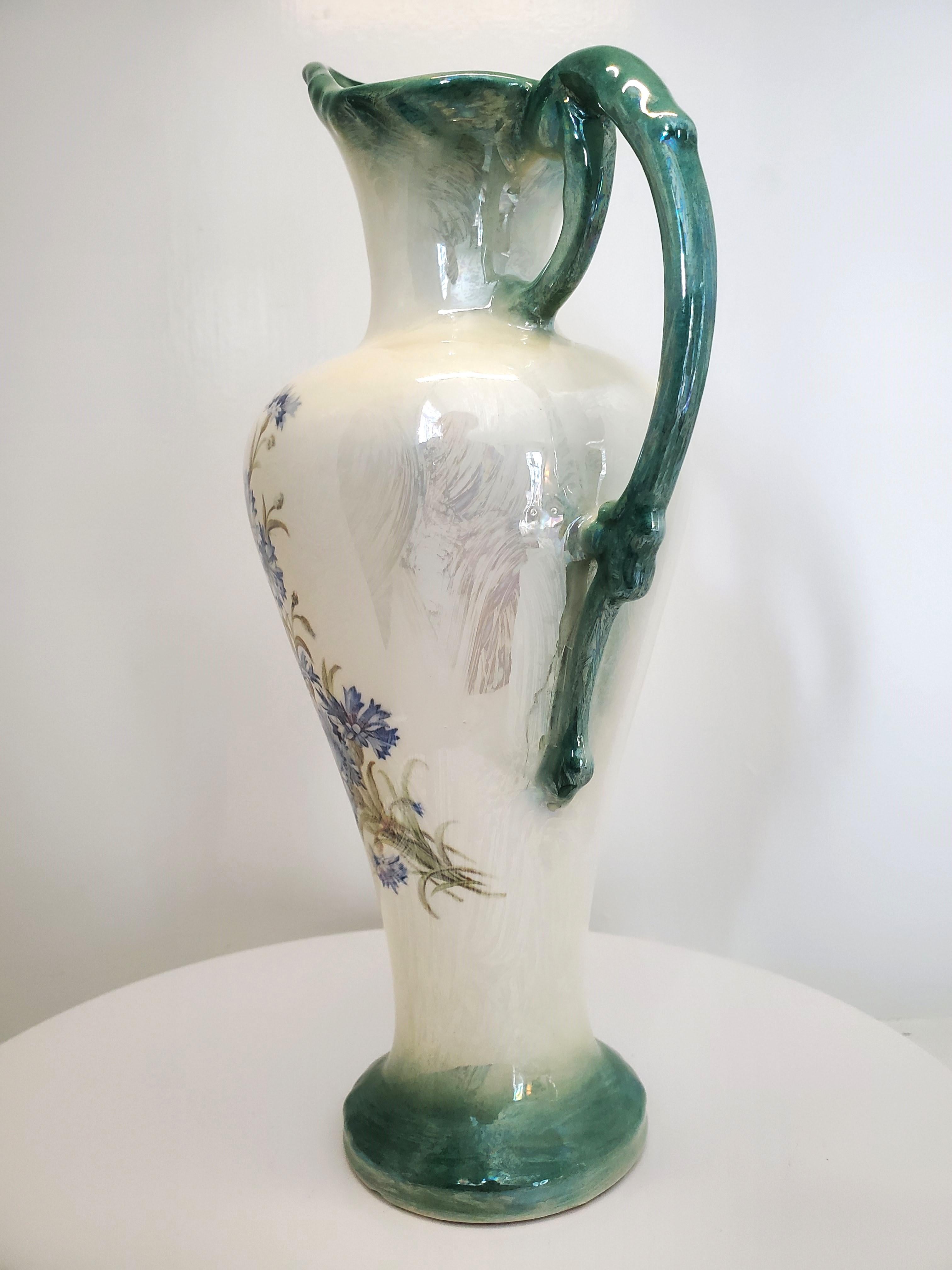 This rare Victorian opalescent lusterware ewer boasts a pearly iridescent base colour and blue-green trim on the mouth, handle, and base. It is decorated with a bouquet of light blue flowers and light olive-green stems, leaves, and buds. The
