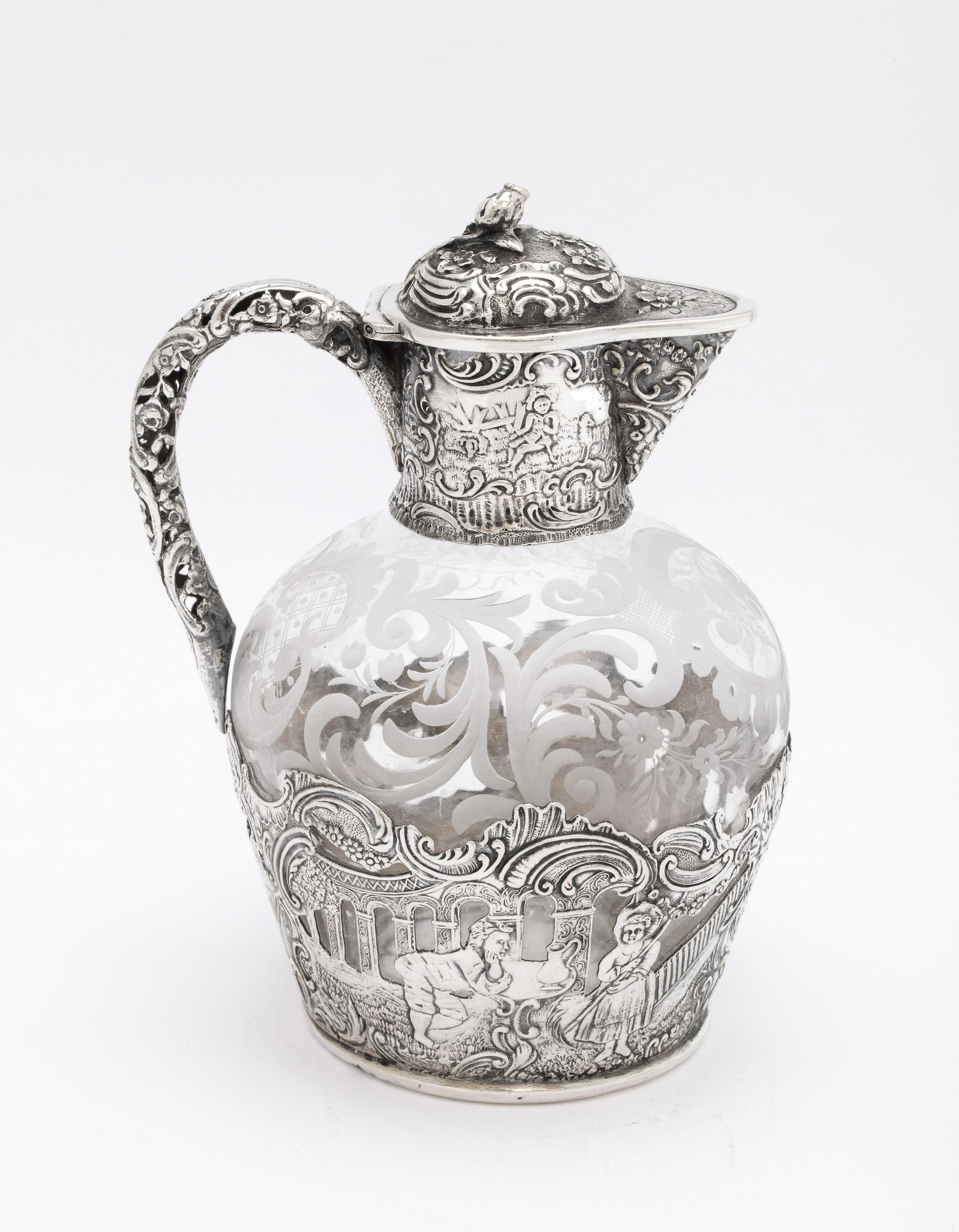 Victorian Period, Continental Silver (.800)-mounted, etched glass liqueur decanter with hinged lid, Hanau, Germany, Ca. 1900. Glass is etched with swirls and flowers; the silver is decorated with country scenes. The handle is pierced. Measures 6