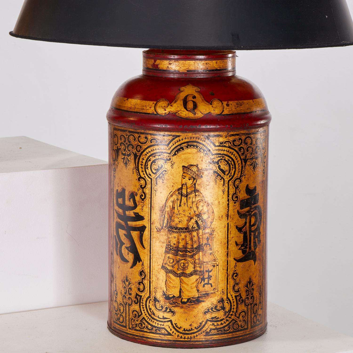 Late 19th c., England, a pair of gilt and red enameled sheet metal tea canister table lamps, decorated with robed Mandarin figures standing by tea boxes and framed by faux-Chinese characters and numbered 5 and 6 respectively, mounted with a brass