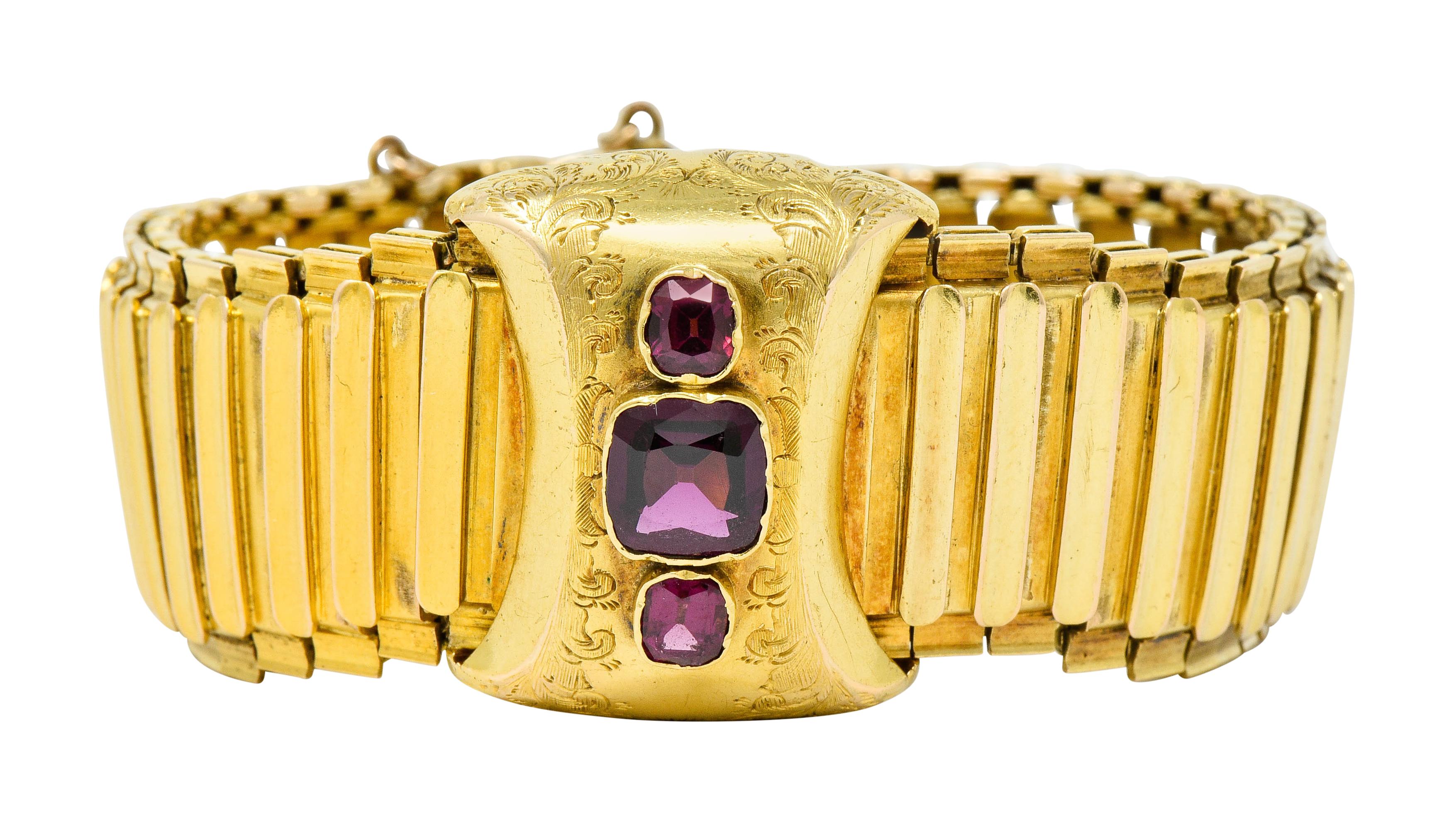 Bracelet is comprised of articulated and deeply ridged rectangular links that flawlessly nest together

With a large hour glass shaped central station, deeply engraved with scrolling foliate

Centering three bezel set rhodolite garnet, cushion cut