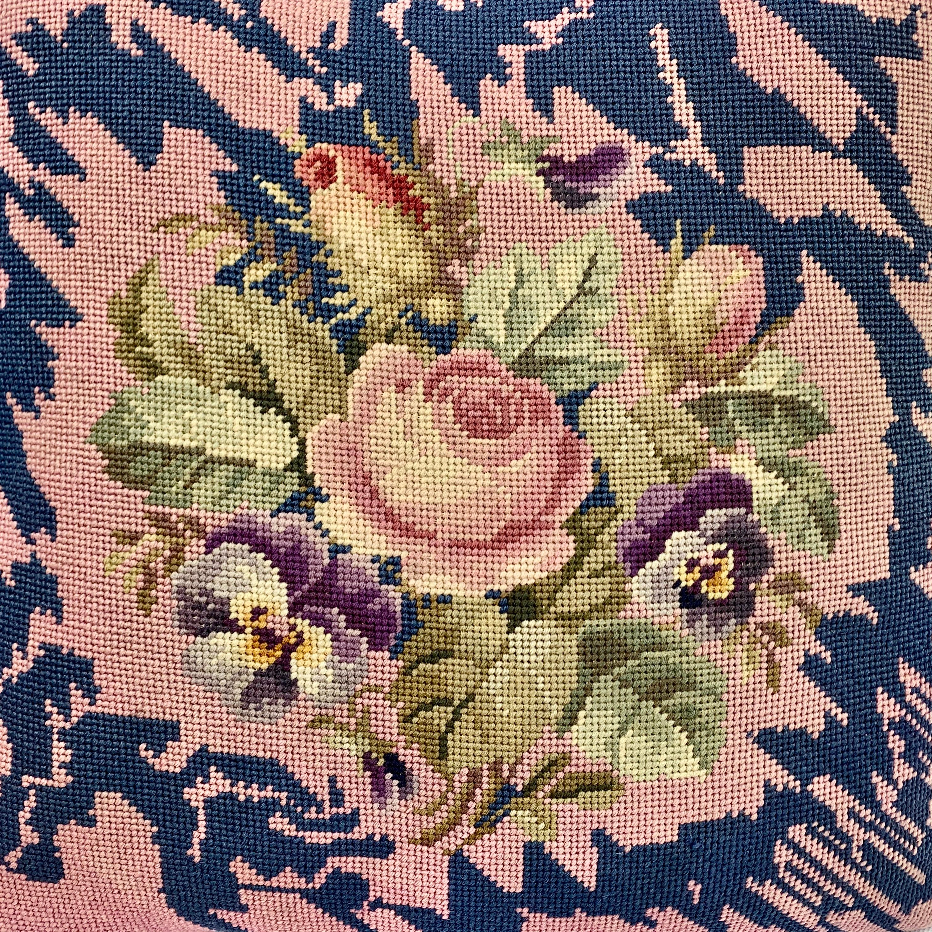 Victorian needlepoint cushion or decorative pillow. The subject is a spray of a large single rose, rose buds, pansies and green leaves. Surrounding the floral spray is a zig zag pattern done in navy blue. The new backing and piping is in a rose