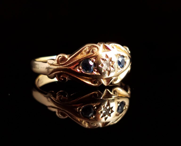A beautiful antique early 20th century 18 karat yellow gold, Sapphire and Diamond ring.

It has a chunky fancy scrolling frame around the ring face with a single gypsy set diamond shouldered by two deep blue sapphires.

The heavy shoulders taper off