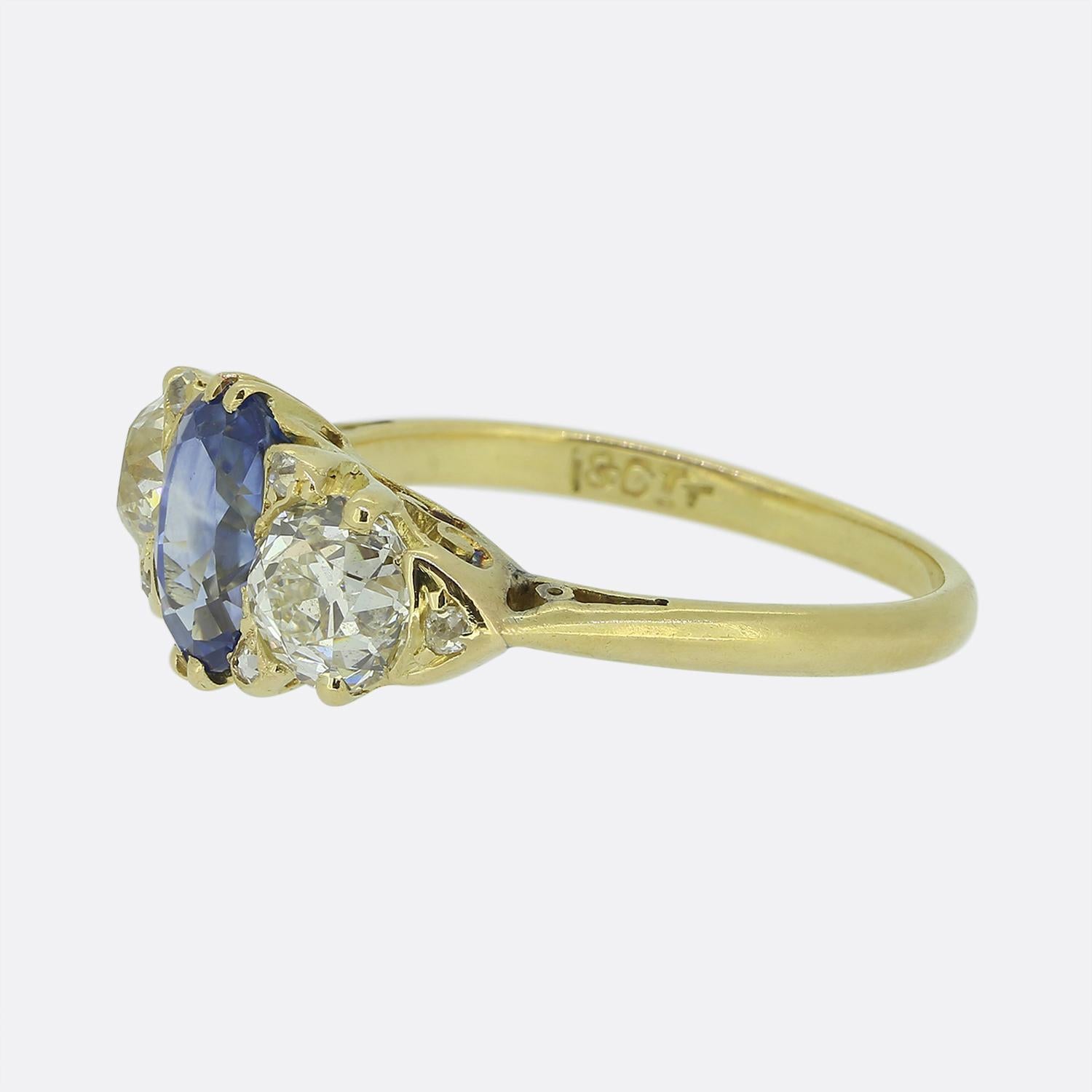 Cette charmante bague à trois pierres est ornée en son centre d'un captivant saphir bleu de taille ovale. Autour de cette pierre principale se trouvent deux diamants blancs taillés dans des mines anciennes, chacun étant étroitement serti afin de
