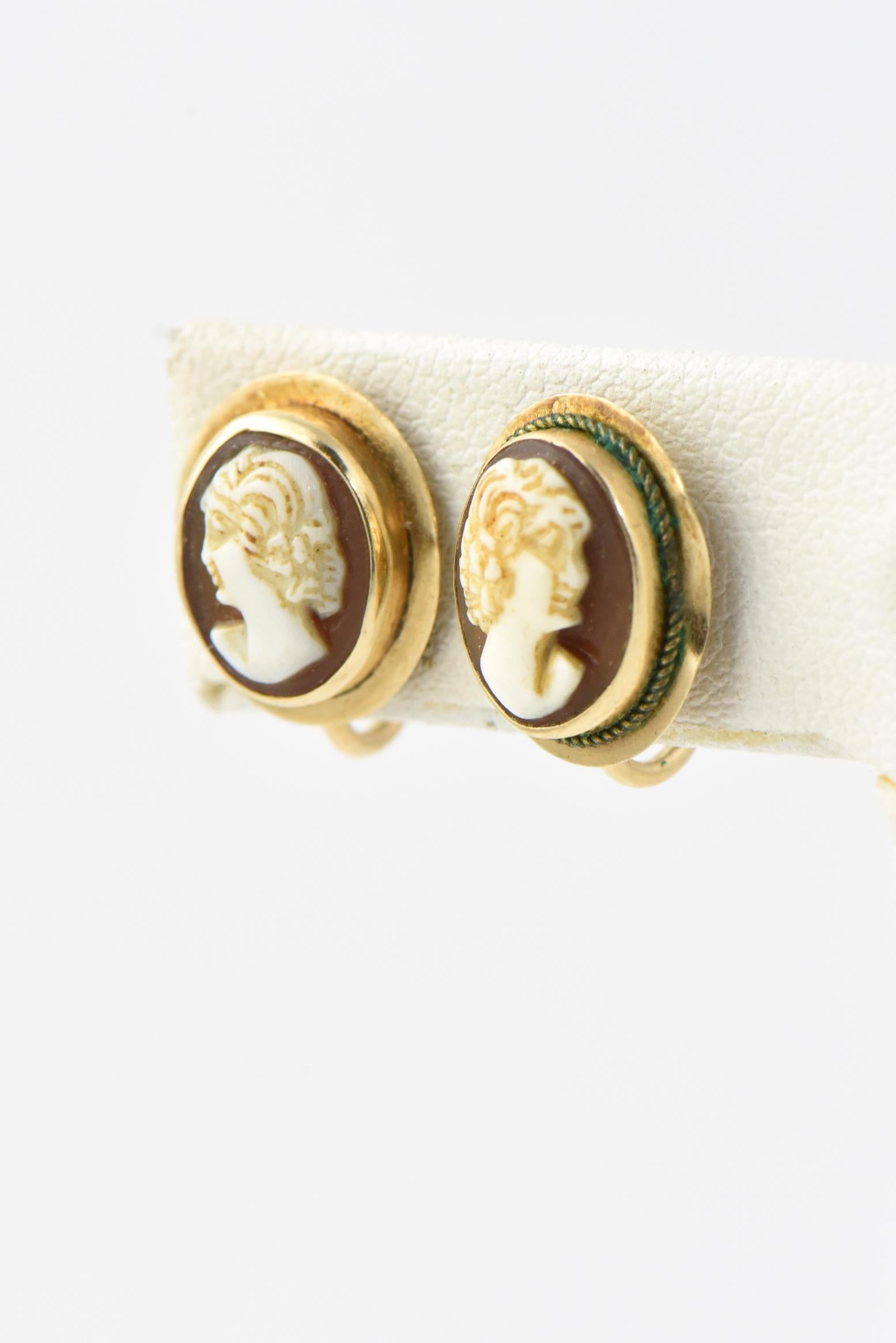 Small Victorian carved shell cameos featuring a portrait of a woman mounted in a raised gold bezel on a gold oval base. These earrings have a traditional Victorian screw back with no post. Marked: 14K. Age wear, tarnished, frame bent.