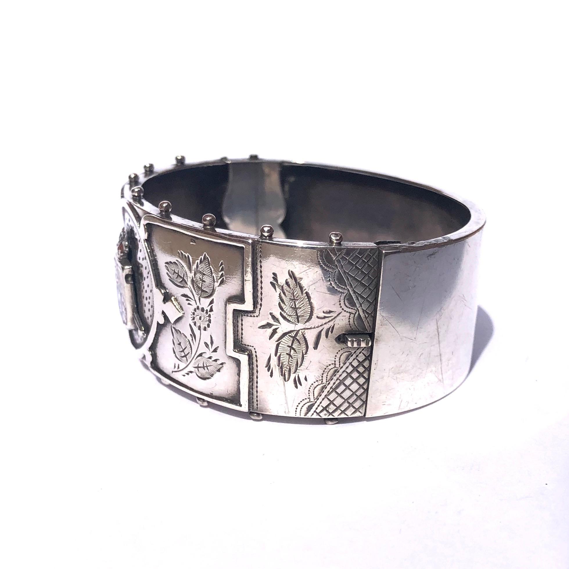 This silver bangle has exquisite engraving and placed at the centre of the front panel is a coat of arms with yellow, red and blue enamel. Around the edge of the front panel there is also beaded detail. 

Inner Diameter: 56mm
Bangle Width: