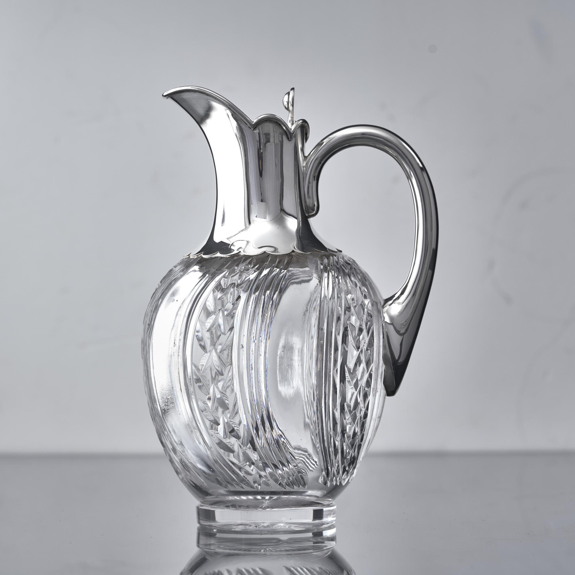 A pretty antique wine jug with round cut-glass body and silver mounts. The swirling bands of fluting and facetting on the glass nicely complement the gentle curves and lobes on the collar. This jug has a hinged lid with cast thumbpiece for opening.