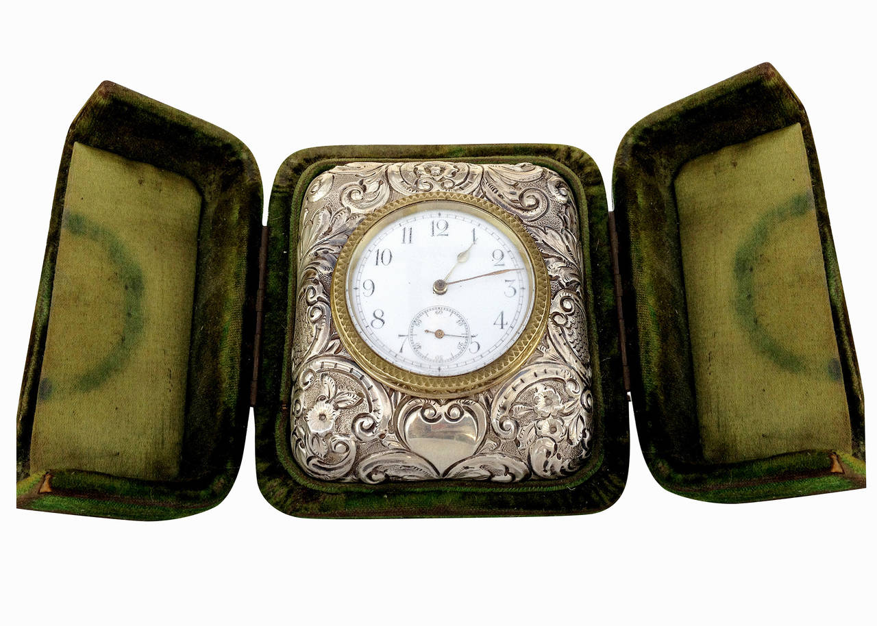 This Victorian silver embossed bedside clock features a brass body clock complete with a brass-cased timepiece and lever escapement along with a solid sterling silver face embossed with decorative flowers. The heavy brass case has an attractive