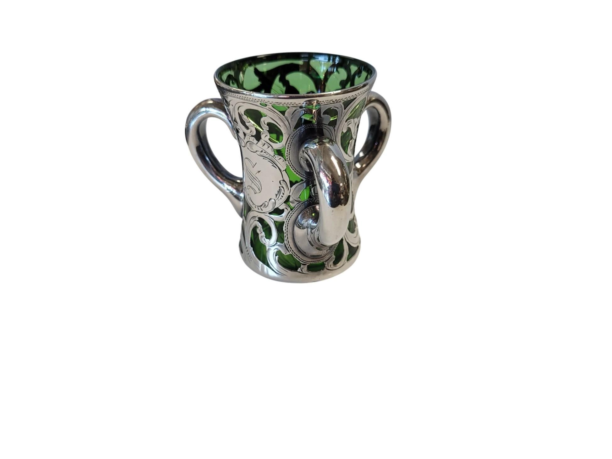 A striking example of late Victorian or early Art Nouveau design, this multi-handled Tyg—also known as a Loving Cup—combines vibrant dark green glass with elaborate silver overlay decoration.

An engraved central cartouche bearing the letter S