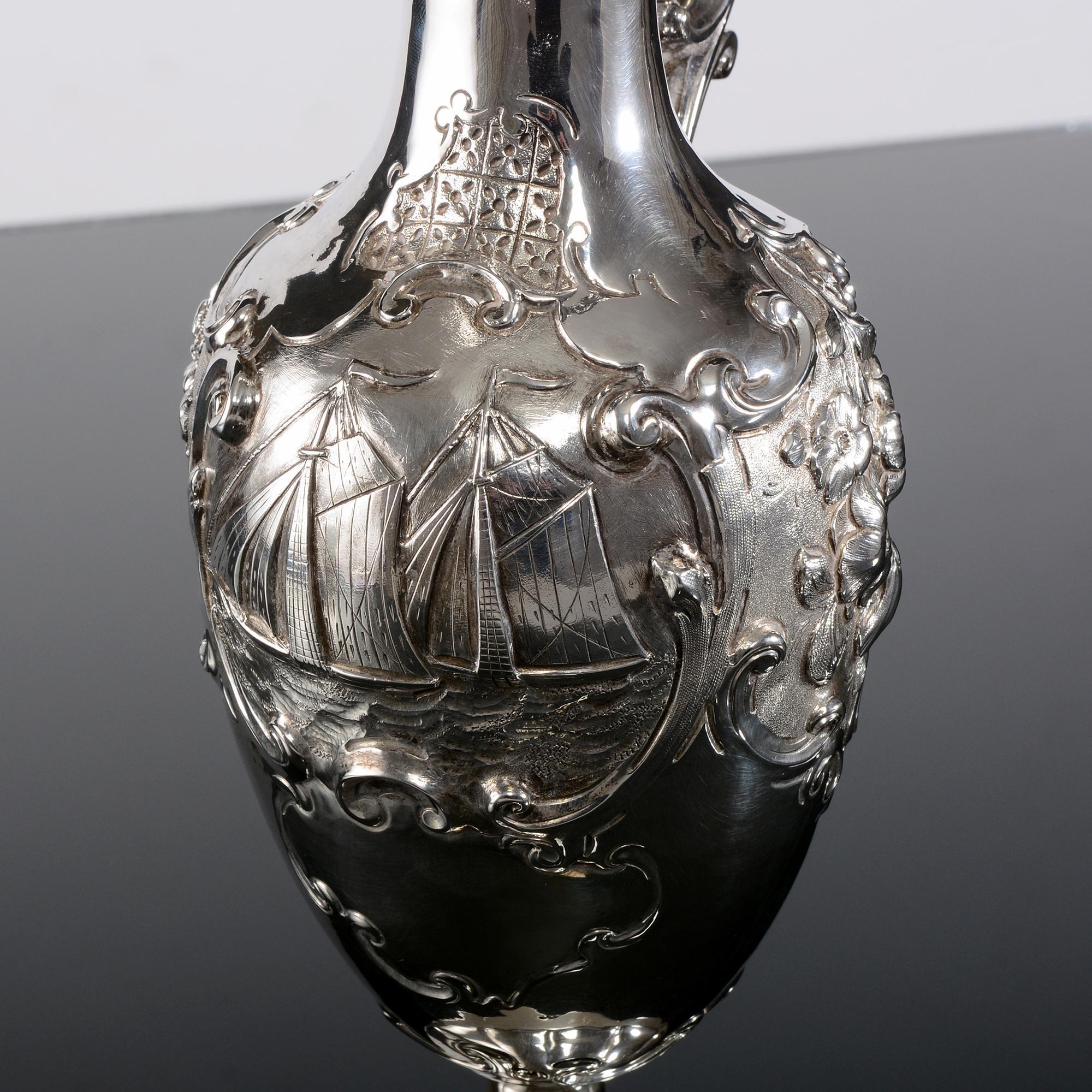 This wonderful Victorian wine ewer is elaborately hand-chased with scroll-framed floral detail on either side of the body and a beautifully detailed sailing scene of two racing yachts. This antique silver jug has a melon style lobed foot, a cast