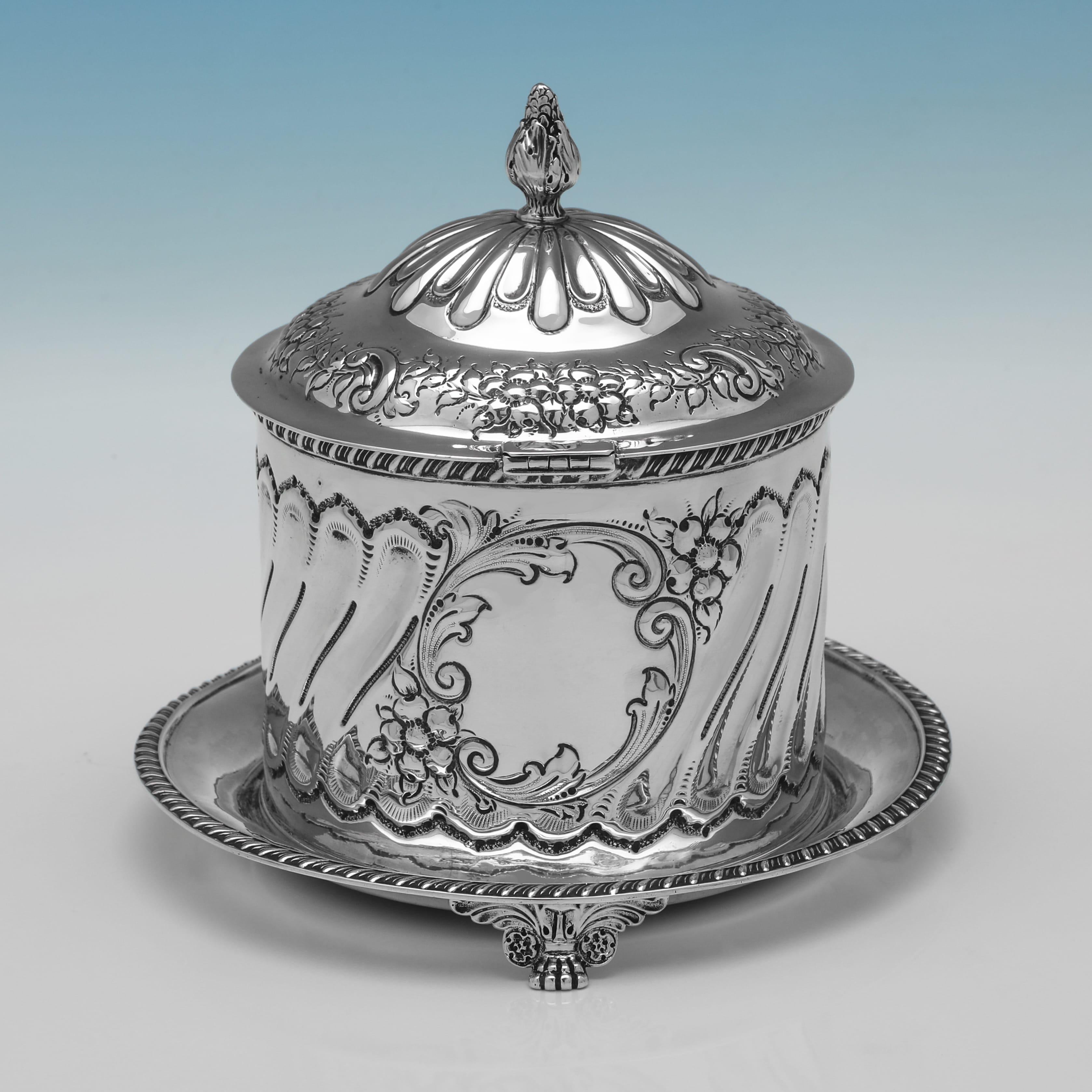 Marchiato a Londra nel 1899 da William Hutton & Sons Ltd., questo ornato portabiscotti vittoriano in argento sterling poggia su un vassoio integrato con piedi a voluta e presenta decorazioni floreali e a voluta, pannelli scanalati, un cartiglio
