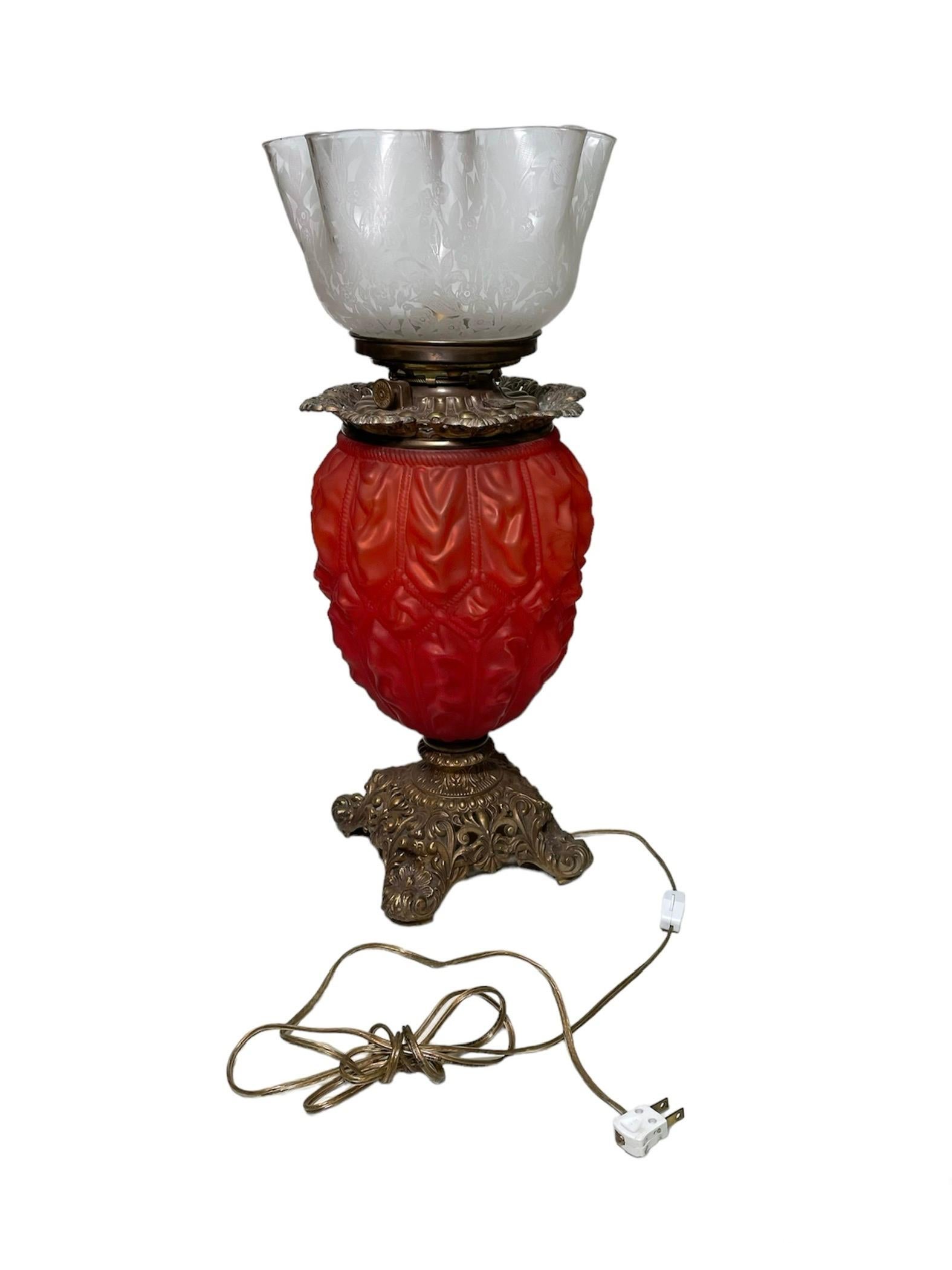 This is a Victorian Brass and Glass table lamp. It depicts a lamp with a large stained red glass bulbous creased body decorated with ropes and supported by a brass square shaped base with four feet. The base is adorned with a repousse of Fleur de