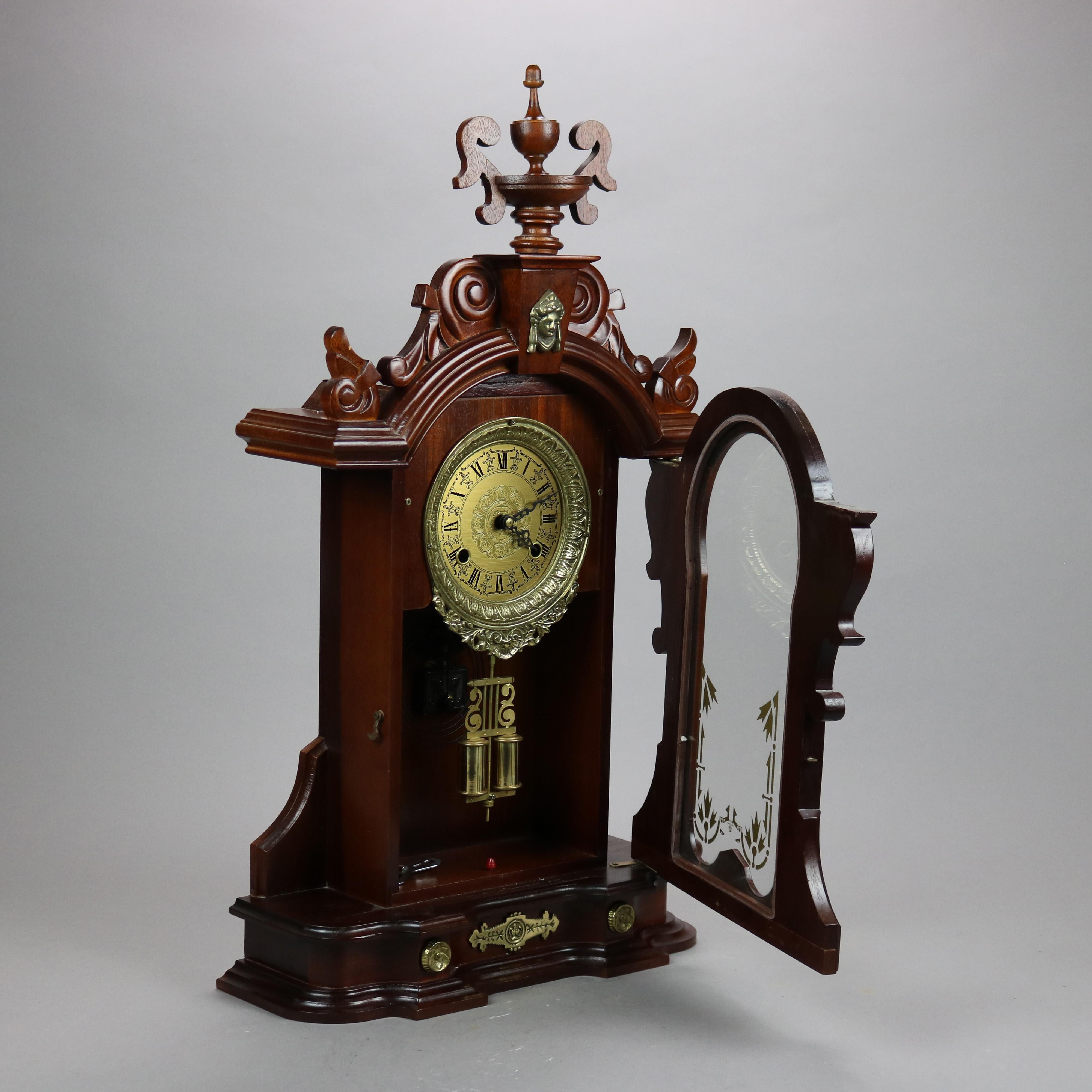 A Victorian style mantle clock offers carved mahogany or walnut case with crest having central urn finial and flanking scroll elements, cast female mask over face having Roman numerals and single glass door with gilt decoration, seated on shaped