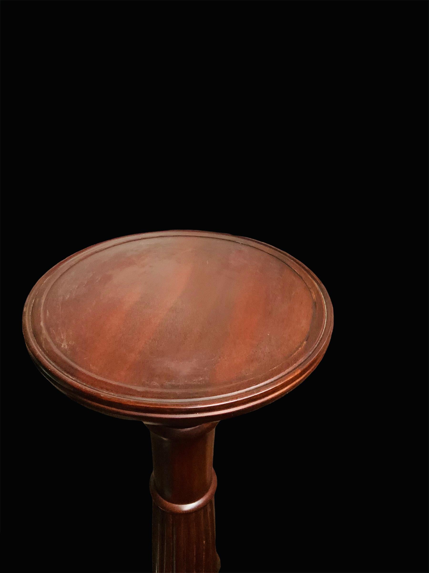 This is a very tall and heavy carved dark solid wood pedestal/torchere/plant stand. It depicts a round top attached to a wide column that is reeded in the center and adorned with the carving of a scalloped petals bud flower with large leaves at the