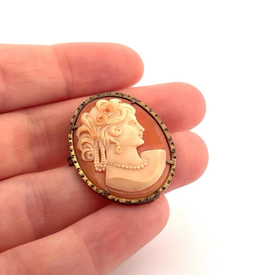 Cette broche camée en coquillage de style victorien présente un portrait de femme de profil, sculpté de façon complexe, avec des boucles détaillées, un décolleté drapé et un rang de perles. Le camée est serti dans une monture en métal mode plaqué