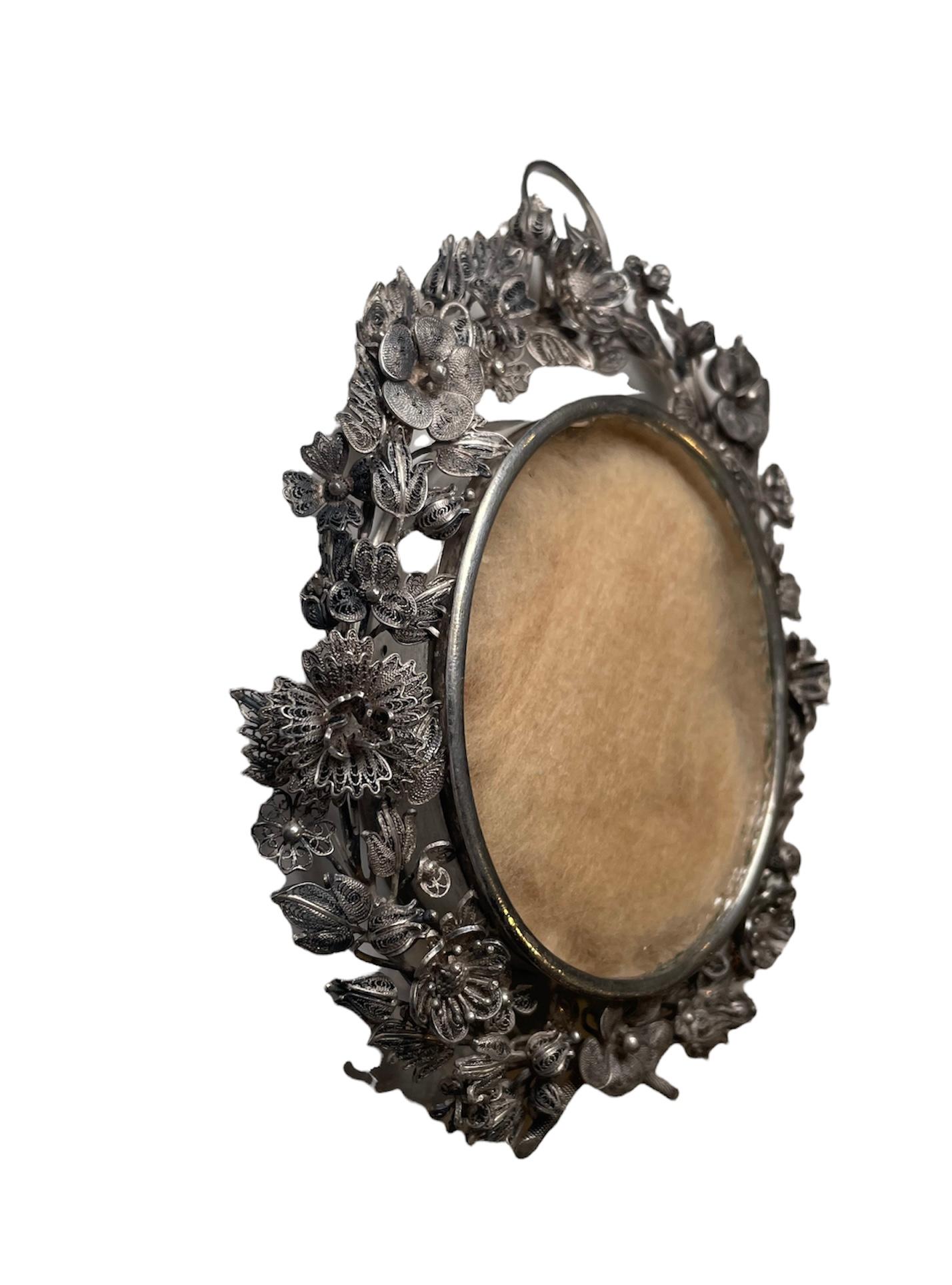 This a large oval silver locket/reliquary pendant. The oval shaped silver case is decorated around its border by different kind of large and small filigree flowers with foliage. There is an oval shaped glass that covers the front and back of the
