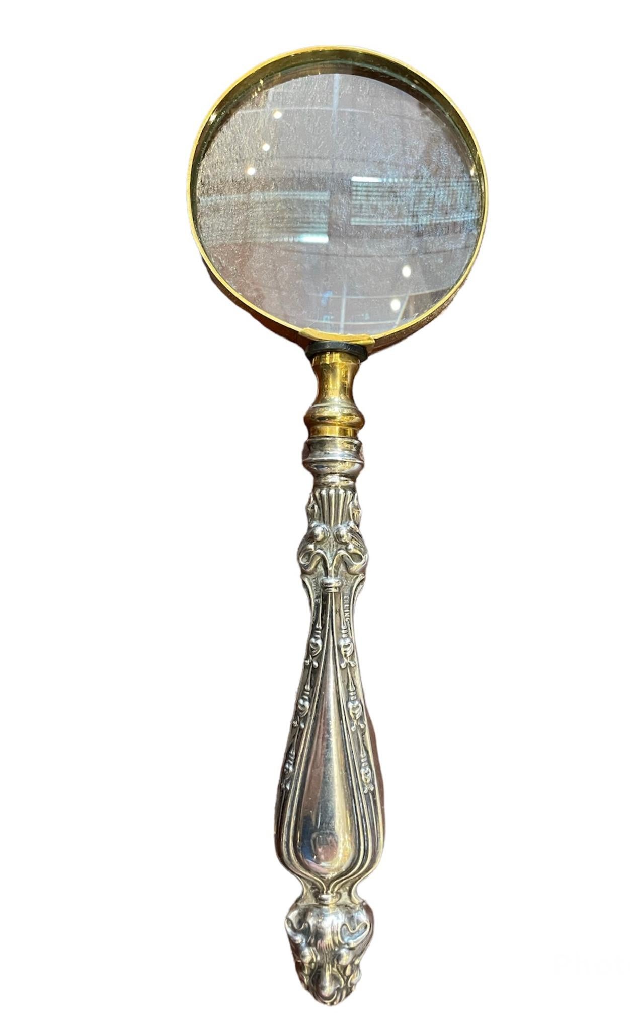 This is a sterling silver and metal magnifying glass. The magnifying glass area is round shaped and framed with a yellow metal. The handle is made of sterling silver and shaped as a drop. It is adorned with the relief of multiple layers of acanthus