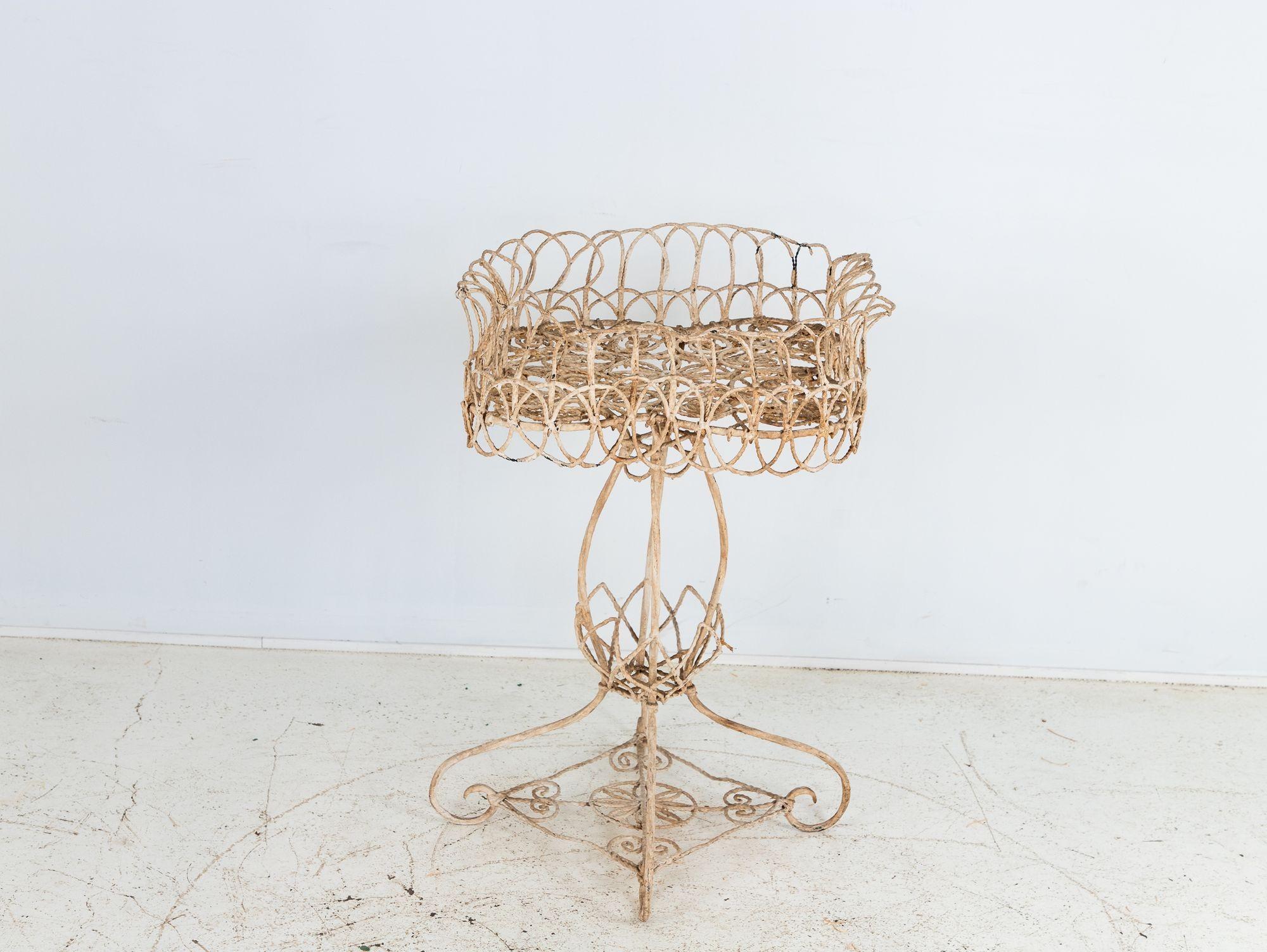 This Victorian-style white-painted wirework plant stand from early 20th-century England features a charming quatrefoil-shaped top elegantly supported by a graceful pedestal base. The design is completed with four beautifully scrolled legs,