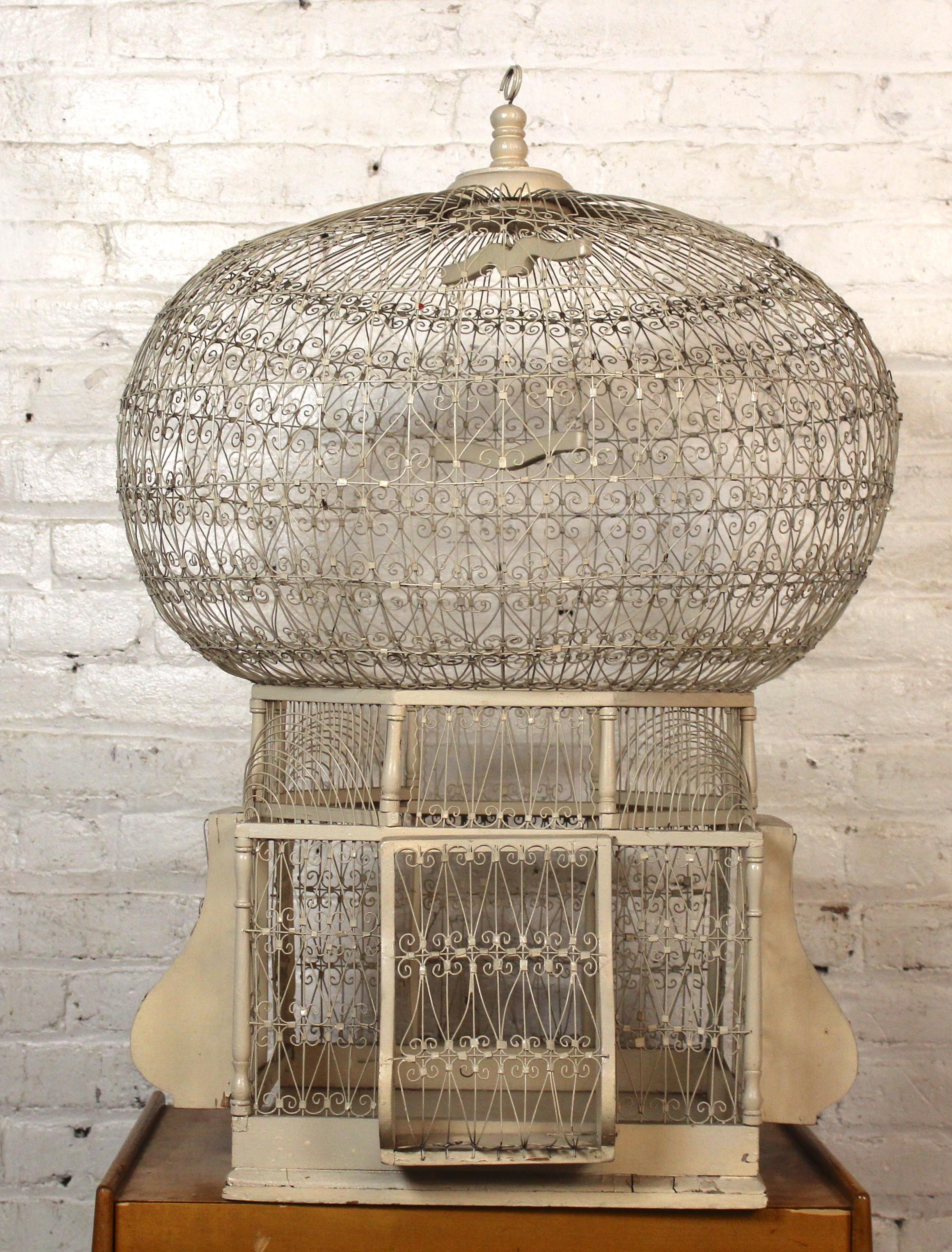 This large antique birdcage features the Victorian style detail, with an intricate wire dome and painted wood frame. Well crafted with scrolled wire and turned wood base. The piece can hang or be placed on a table. The white paint has aged and now