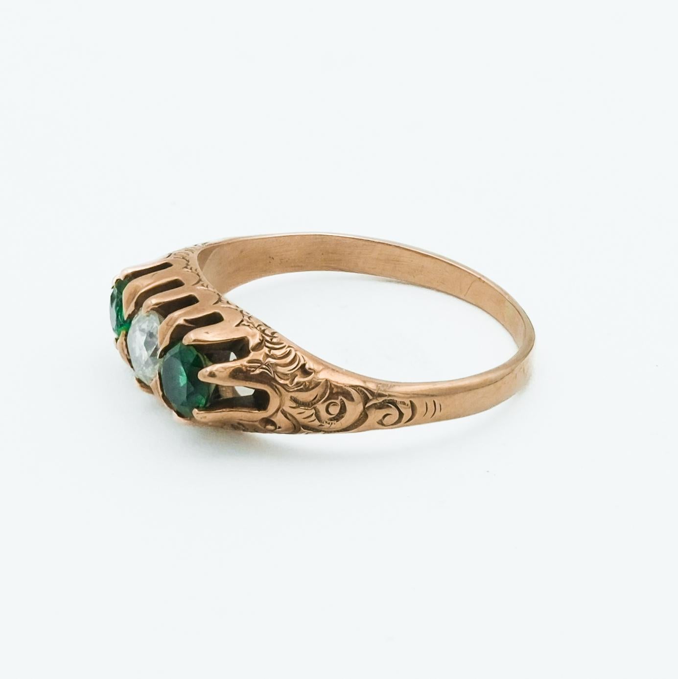 This 10 karat rose gold Victorian ring features a timeless three-stone design with an a diamond center in between two emeralds, a popular configuration during the Victorian era. The emeralds, known for its vibrant green color, symbolizes rebirth and