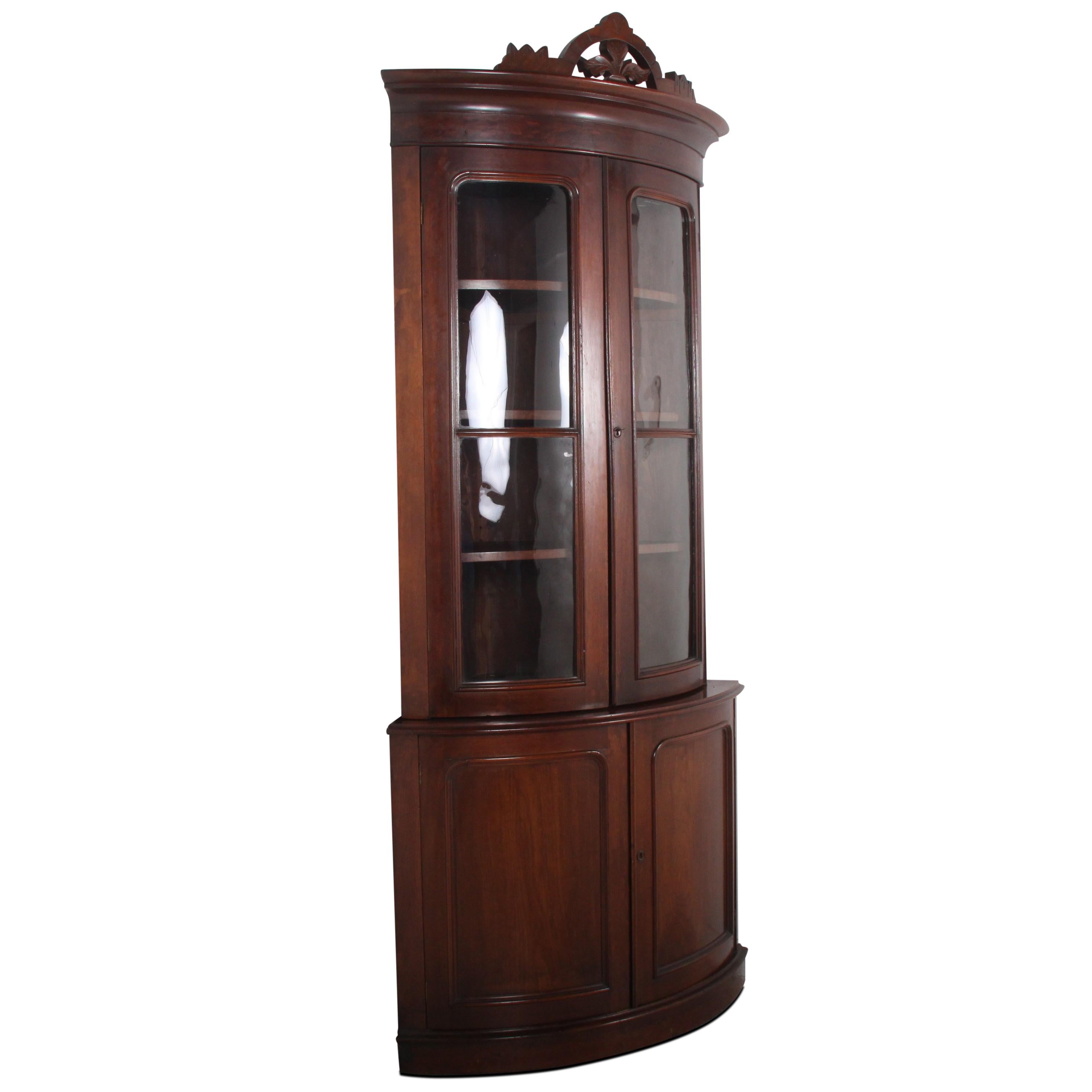 A Victorian bow-front corner cabinet in walnut, the top with three shelves behind original curved glass doors and with lower storage behind a pair of curved- panelled doors. Original carved accent piece on crown is removable if the buyer prefers a
