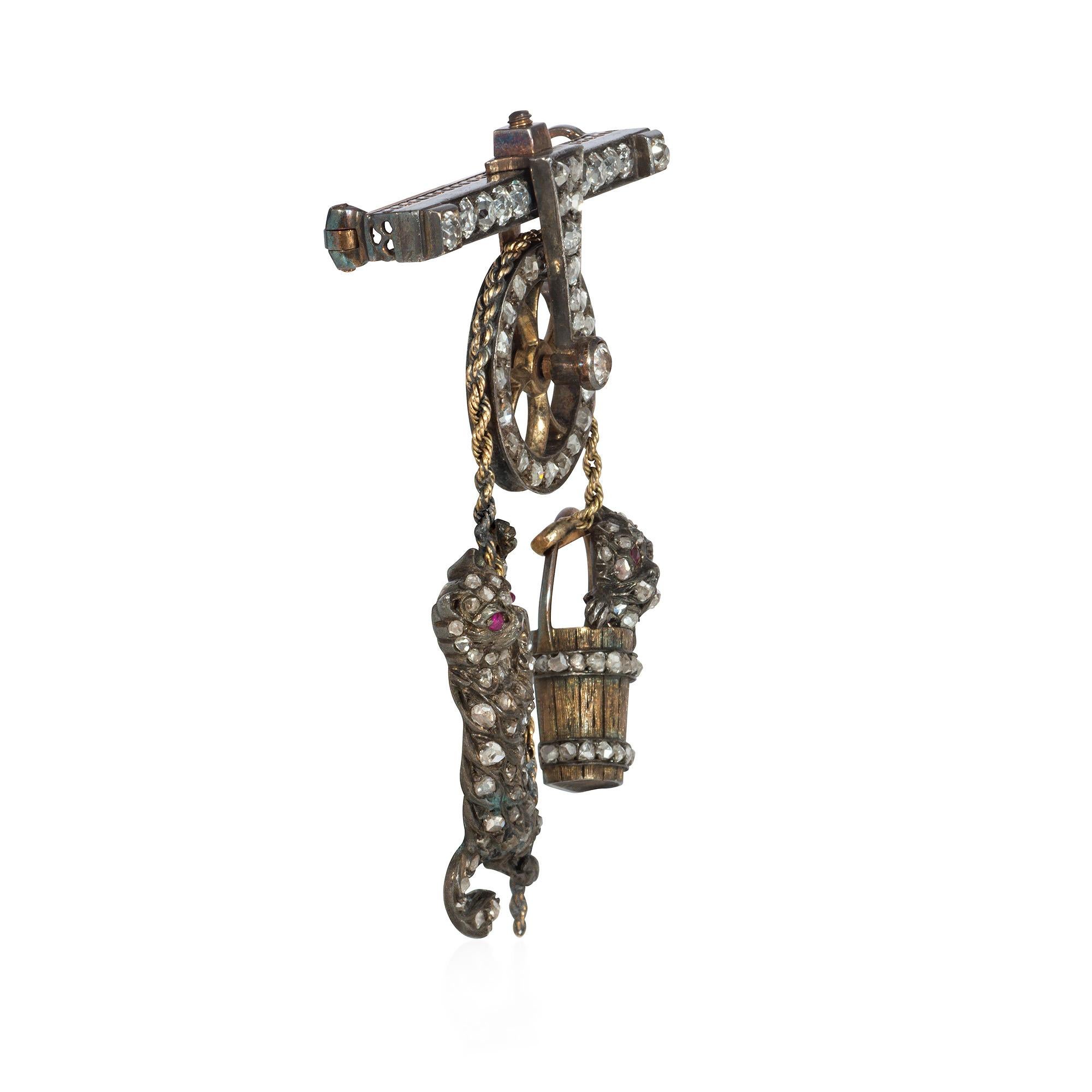 A Victorian diamond brooch in the form of an articulated mechanical pulley suspending a mischevious cat with ruby eyes toying with a dog in a bucket, in sterling silver and 15k gold. England