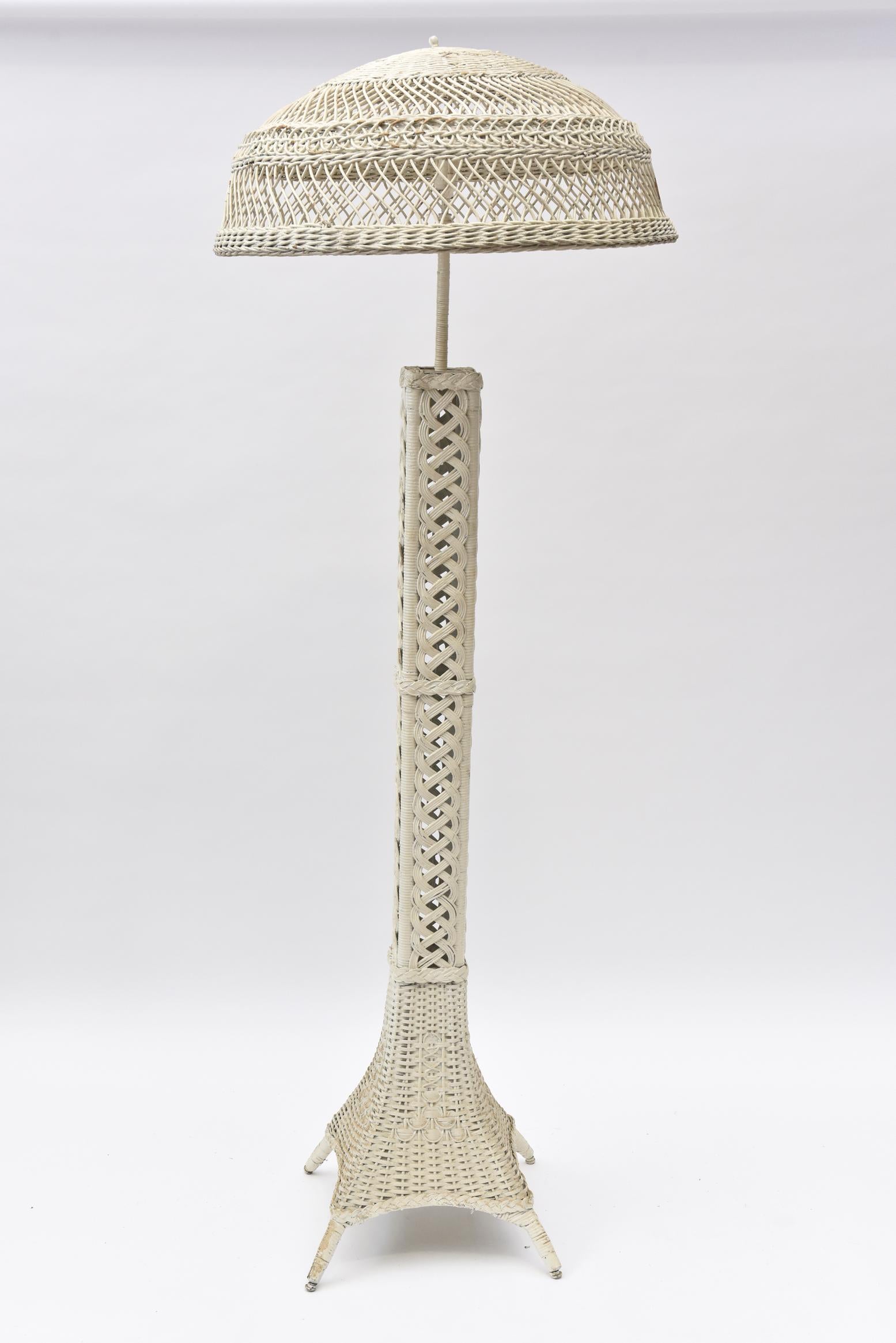 Tall and elegant, this wicker floor lamp has a woven splay foot base with a fancy woven square column leading up to an open weave round dome shade. There is a woven pattern centered in each of the four sides of the wicker base. The electrical cord