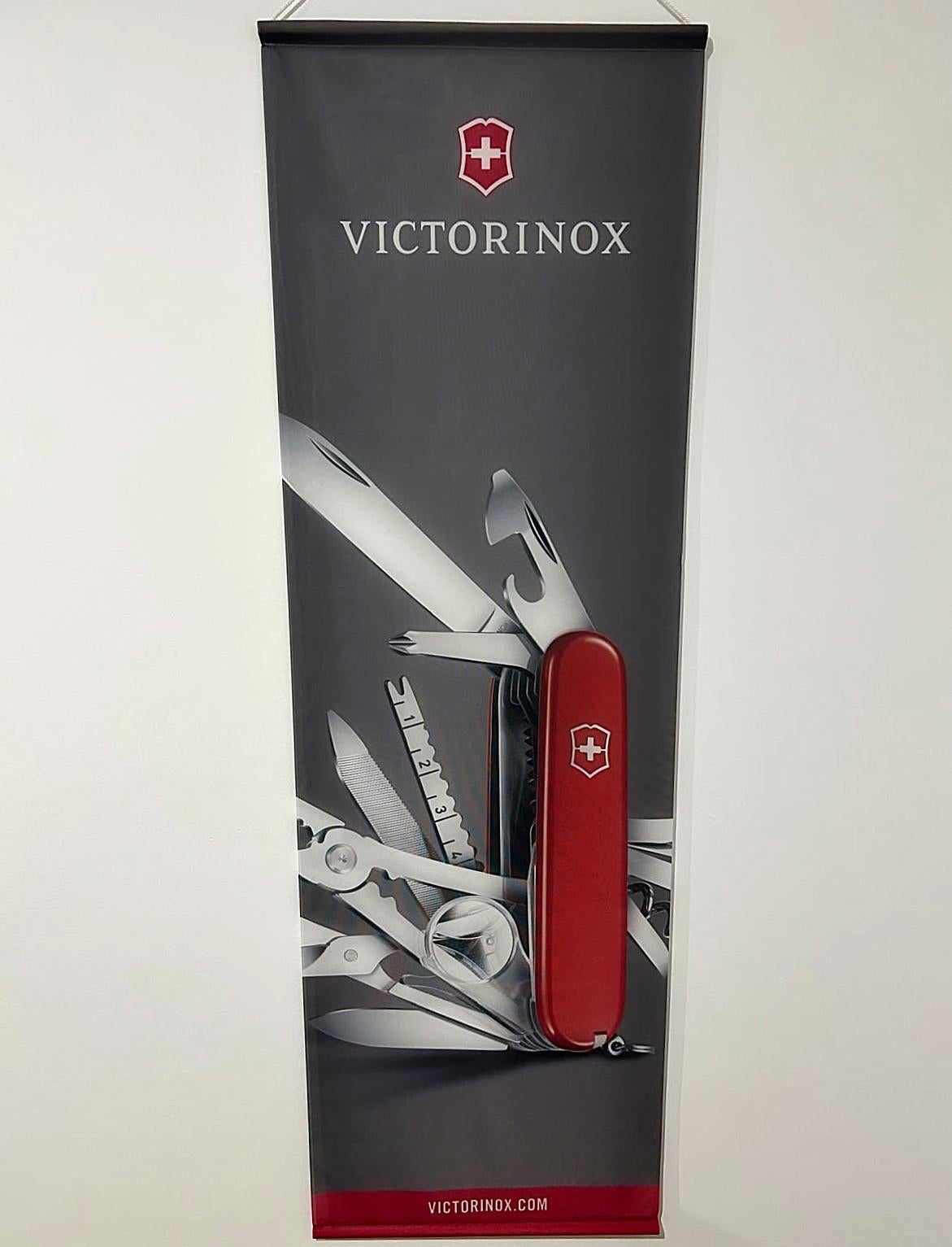 Original advertising banner from the iconic Swiss brand Victorinox, renowned for its precision multifunction knives. This large-format piece is printed on fabric, with black rigid bars at the top and bottom for vertical display. The graphic features