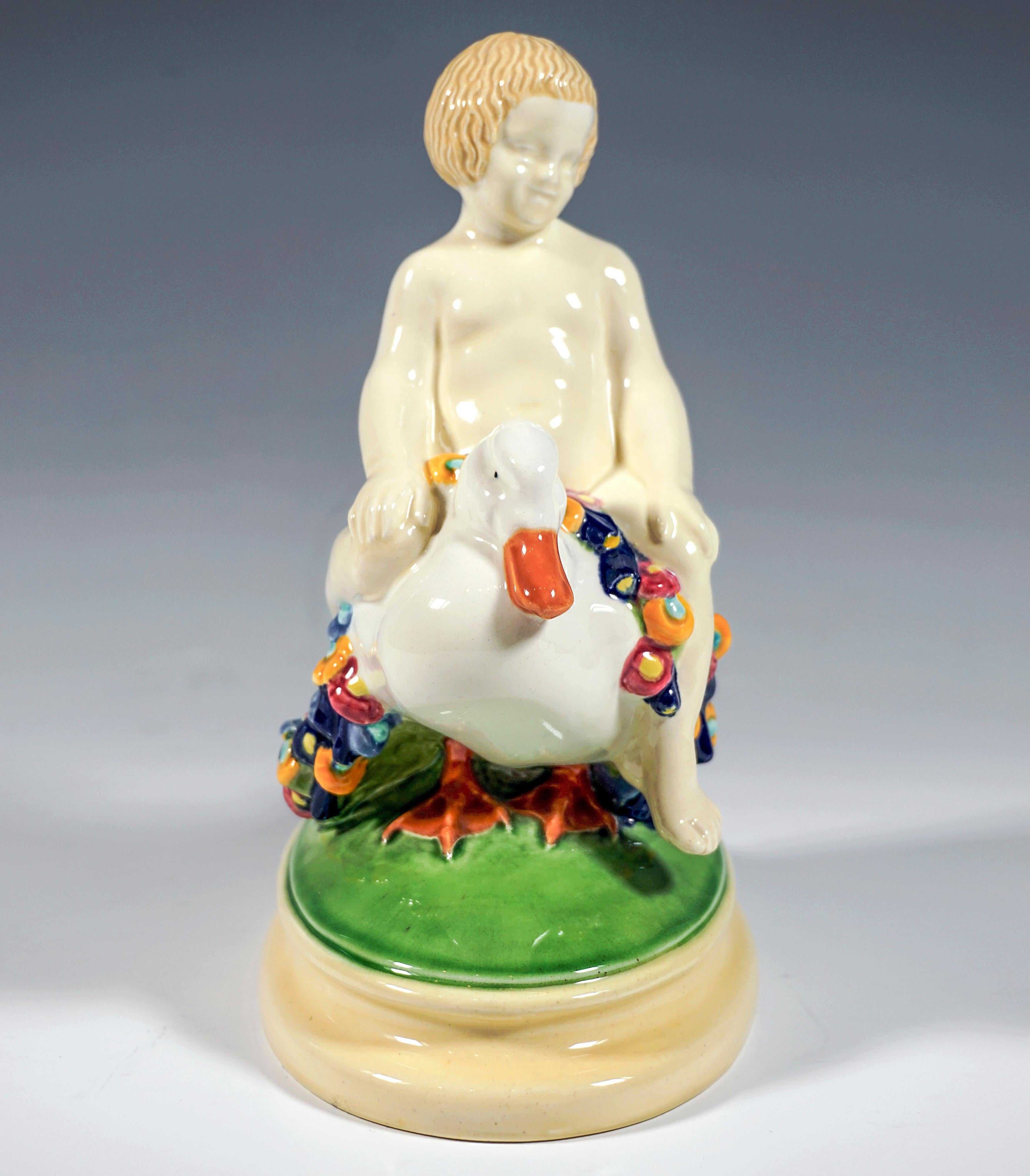 Ausgezeichnetes Wiener Jugendstil-Keramikstück:
Ein blonder Junge sitzt rittlings auf einer mit bunten Blumengirlanden geschmückten Ente und stützt seine Hände auf die Knie.
Die Gruppe steht auf einem ovalen, grün und cremefarben bemalten,