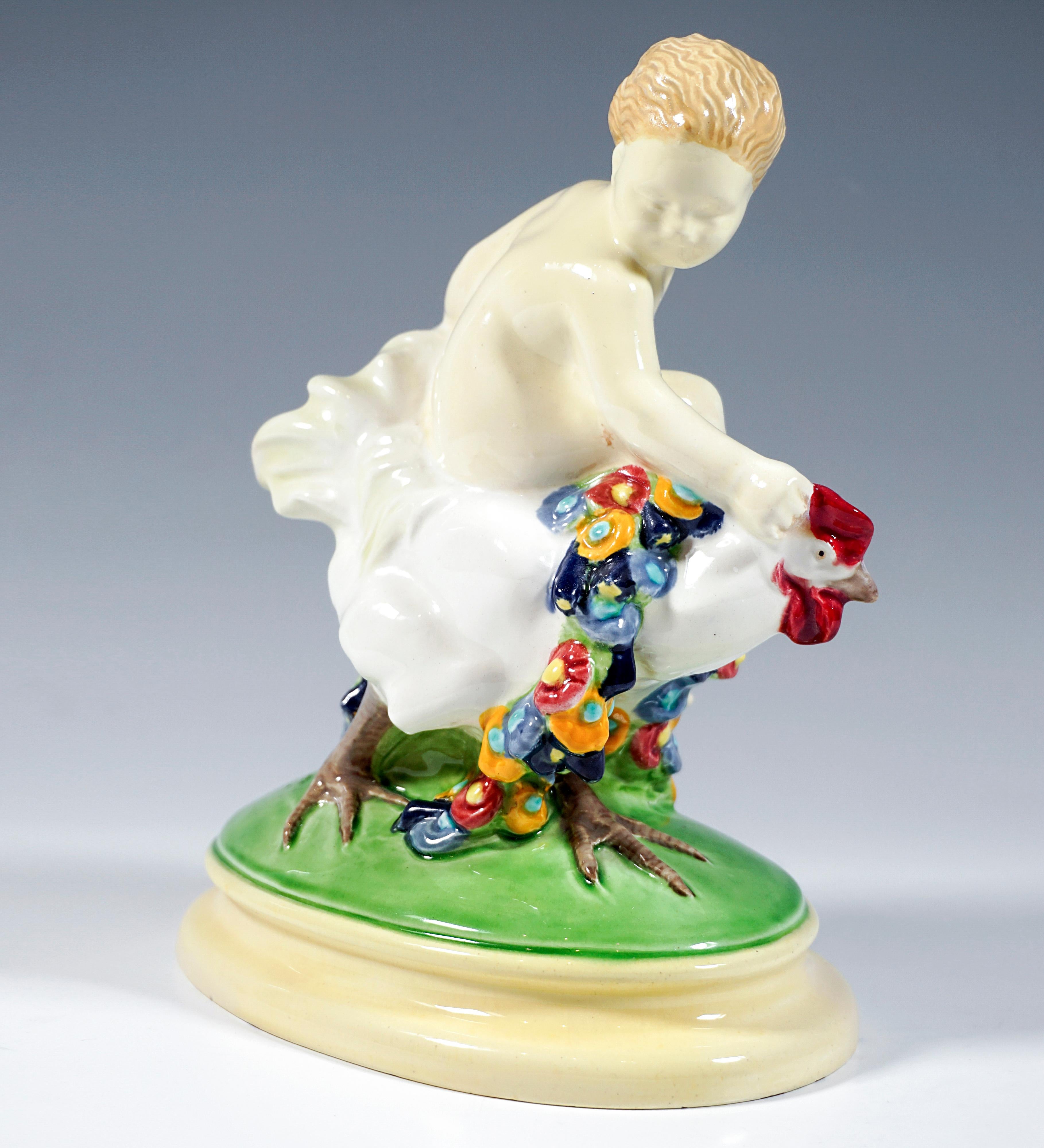 Ausgezeichnetes Wiener Jugendstil-Keramikstück:
Blonder Junge sitzt seitlich auf einem mit bunten Blumengirlanden geschmückten Hahn und hält sich an dessen Kamm und Gefieder fest.
Die Gruppe steht auf einem ovalen, grün und cremefarben bemalten,
