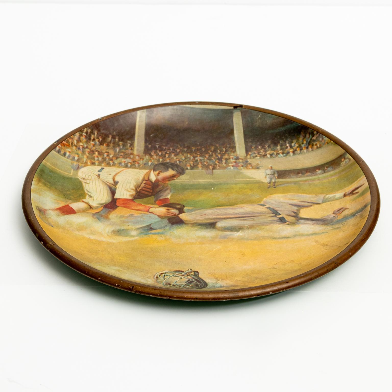 Circa 1920s collectible 10.00 Inch diameter Vienna tin art plate depicting a baseball game with a player sliding into base with the catcher. In the image's background is a stadium of fans wearing hats consistent with the 1920s era. There are no