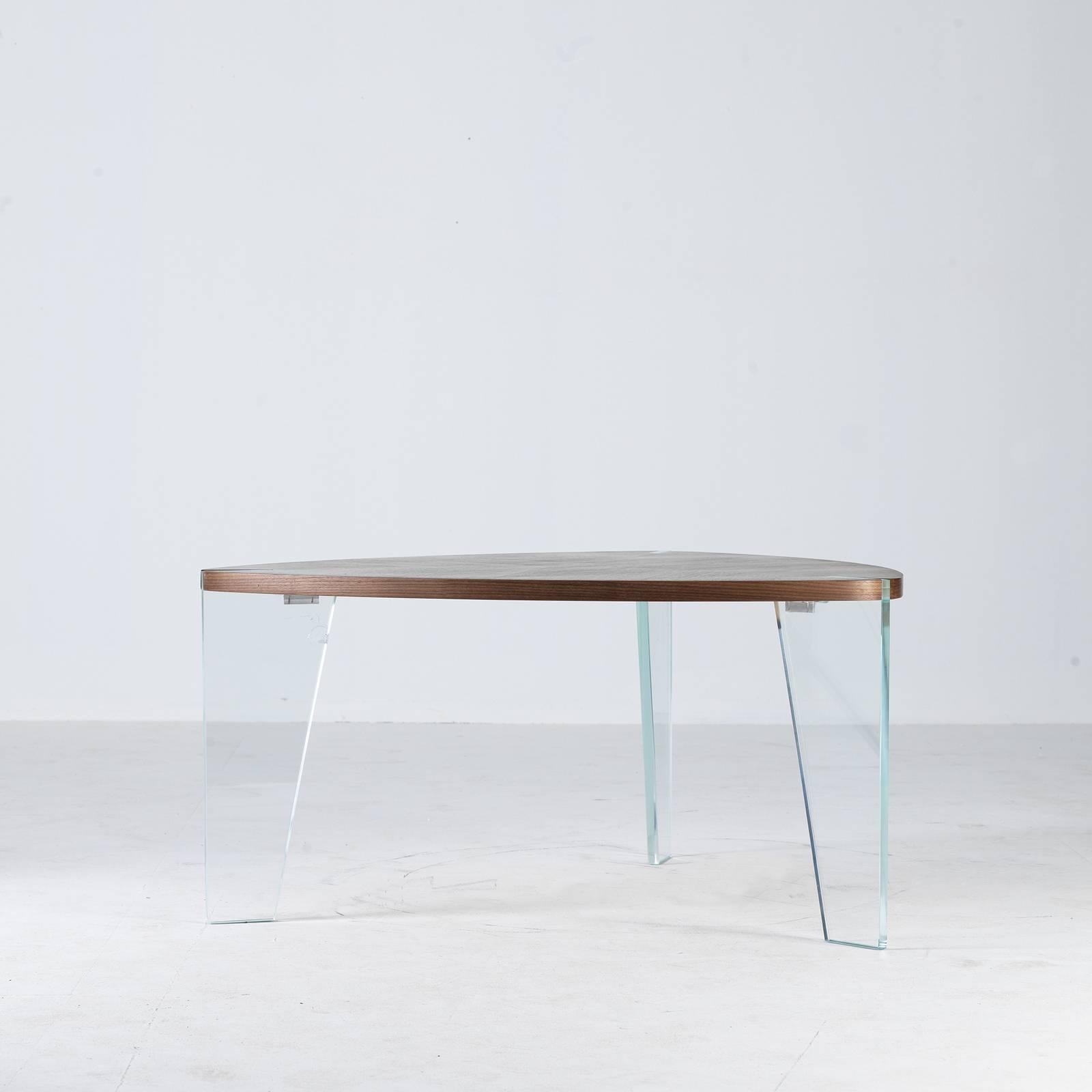 Subverting conventional design norms, this striking coffee table is composed of three curved extra-clear tempered glass legs framing a drop-shaped walnut top with an interlocking joint. The herringbone pattern of the wooden shelf creates an