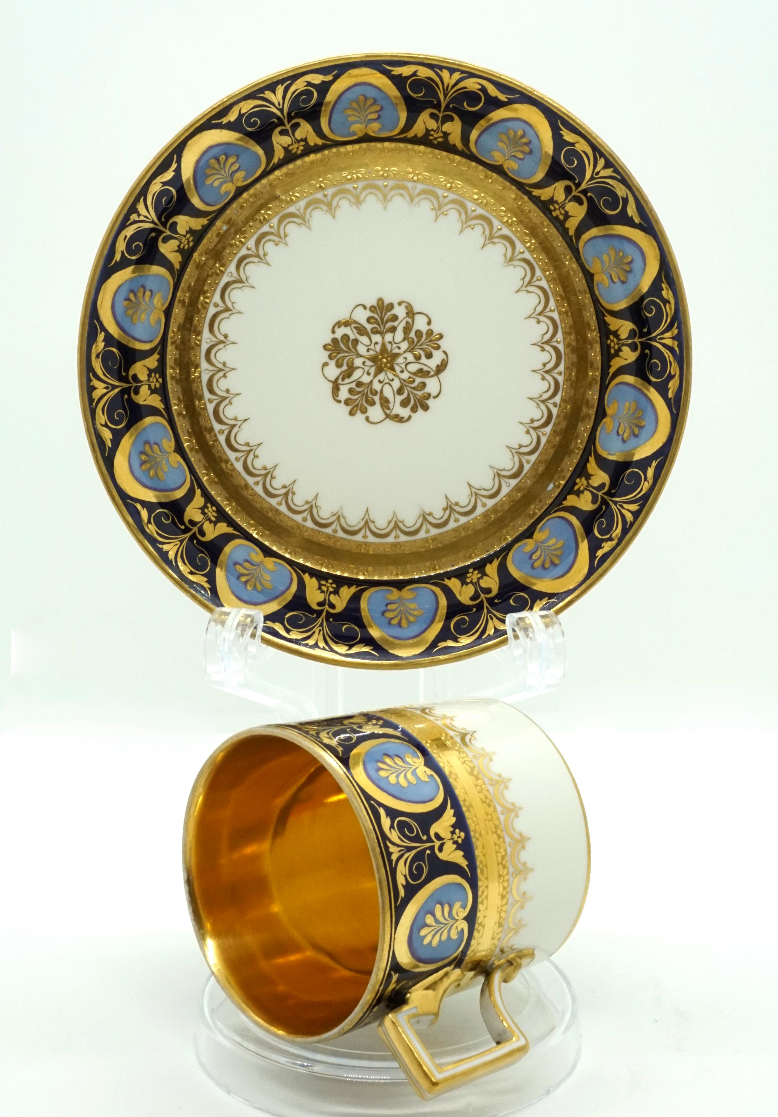 The upper area of the cylindrical white cup has a cobalt blue band with light blue medallions, which are leaf-shaped framed with gold and painted with leaf panicles, with repeated golden leaf and tendril motifs in between. Double gold rim on the