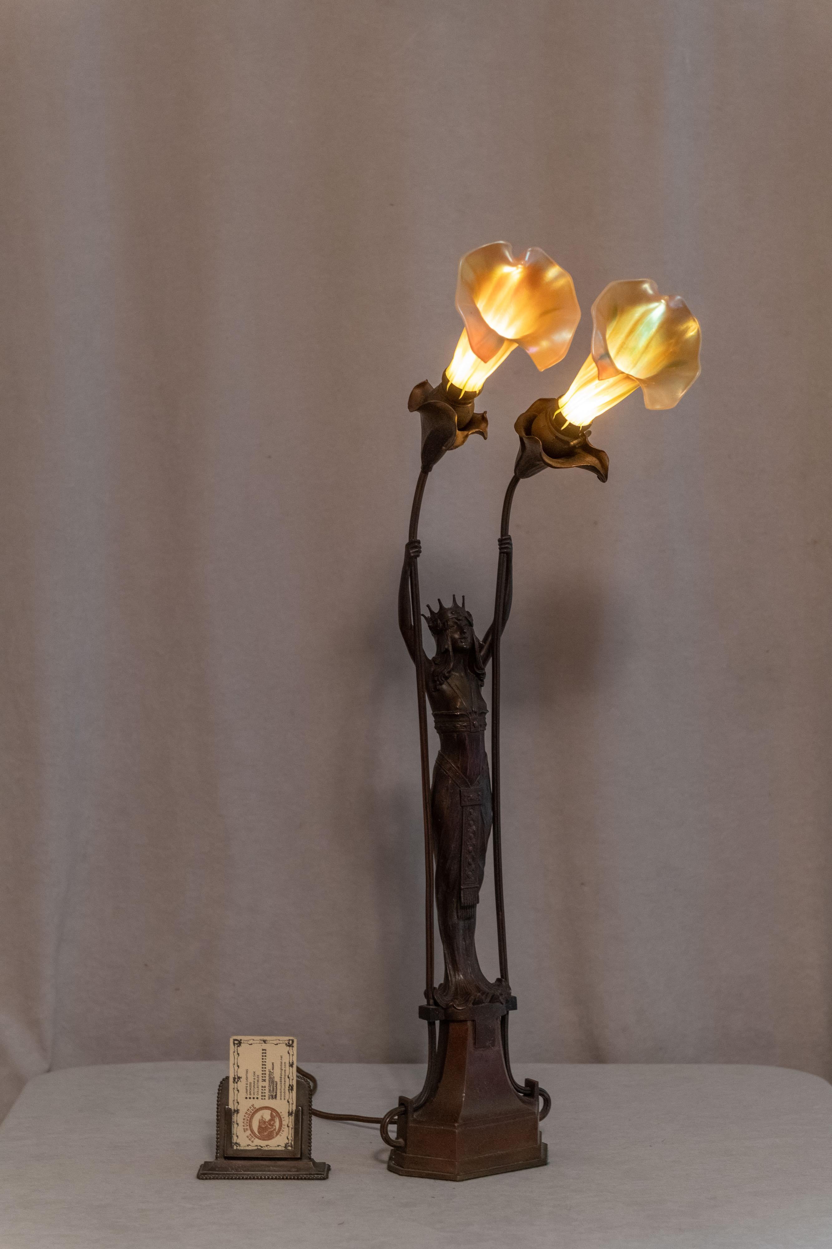  On y trouve des lignes très épurées et un engagement envers le mouvement sécessionniste viennois. Notre jeune femme porte une couronne et tient deux abat-jour en verre d'art à plumes tirées. Les abat-jour sont des pièces de rechange, mais ils
