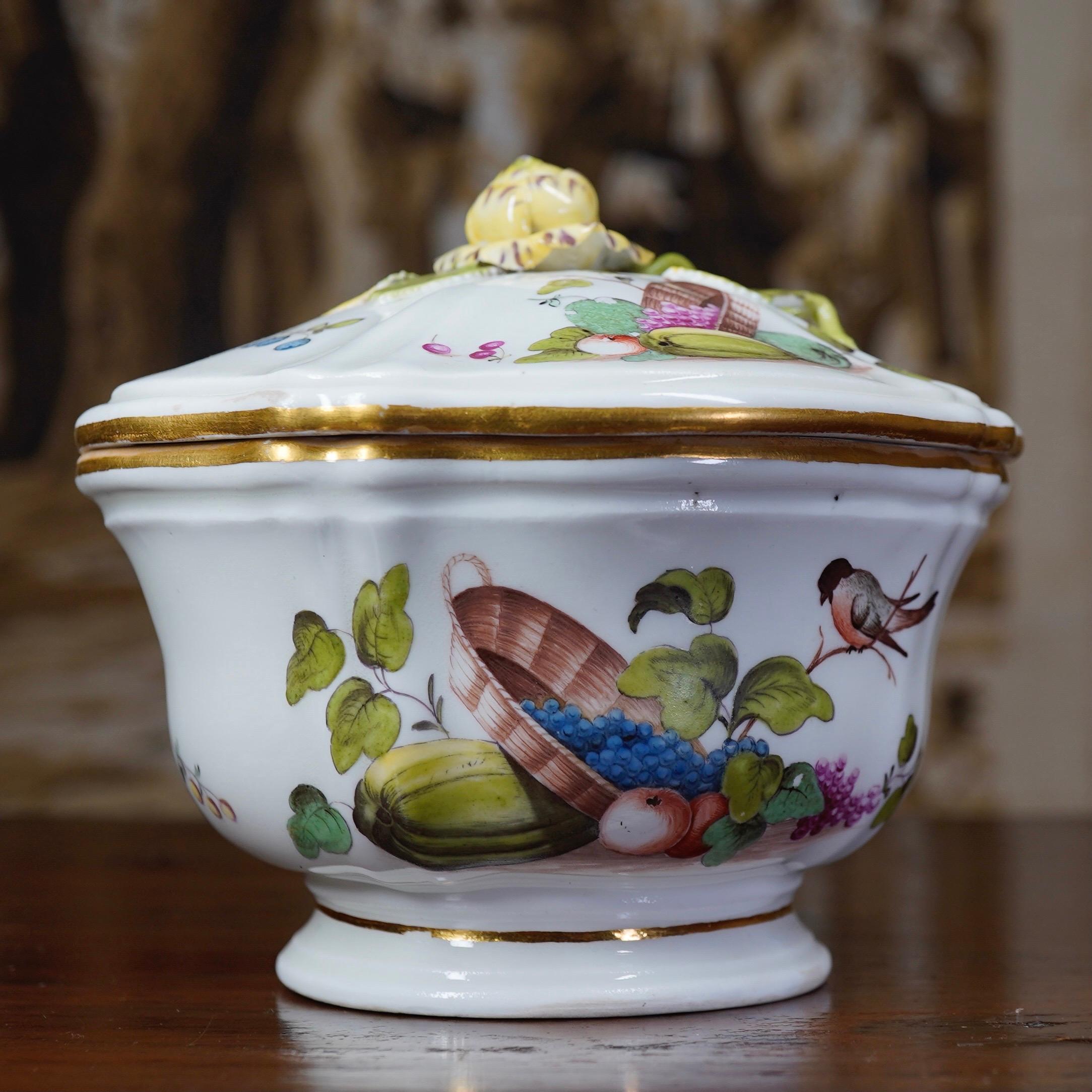 Vienna sucrier of elaborate rococo form, with ribbed body after a metal form, painted with a wicker basket spilling grapes and apples, resting on a melon, with a bird perched to one side, the reverse with cut melons and grapes, the lid with similar