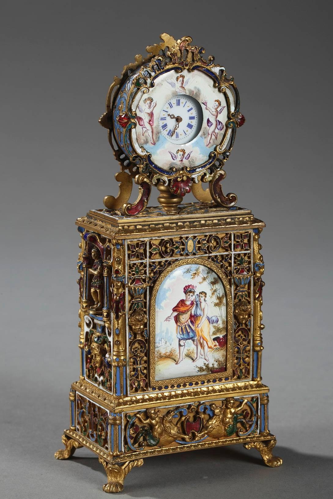 Small table clock in gilt brass richly decorated with openwork scrollwork, acanthus leaves, flowers, and masks in multicolored enamel. The main faces of the base are ornamented with two enamel plates that represent mythological scenes. The side