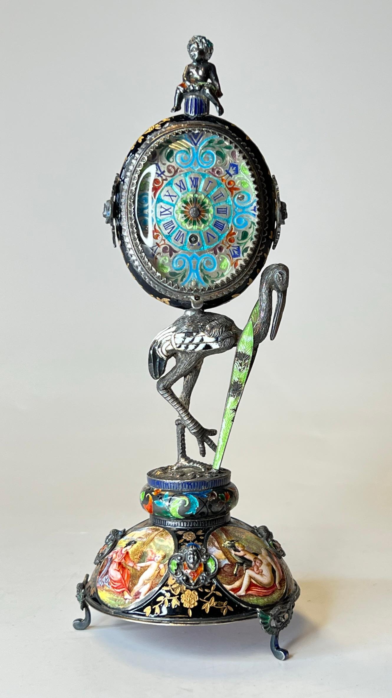 Austrian enameled table clock, attributed to Hermann Bohm, with stork standing on domed base raised on winged cherub feet, supporting an oval shaped case decorated with painted cartouches depicting classical mythological figures.