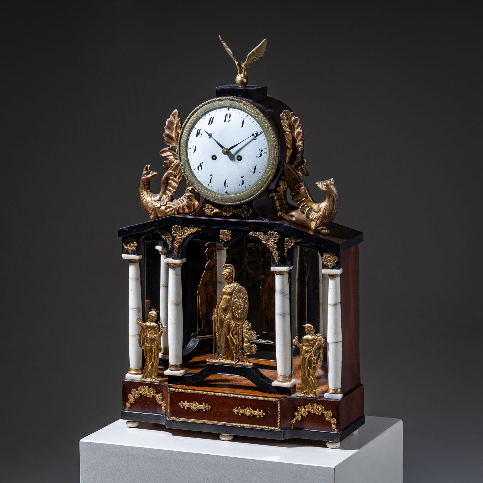 An elegant Viennese portal clock resting on a rectangular mahogany base. The ebonized case is richly adorned with fire-gilded bronze appliqués. Flanking the clock are carved decorations depicting chimerae with eagle heads, and it is crowned with