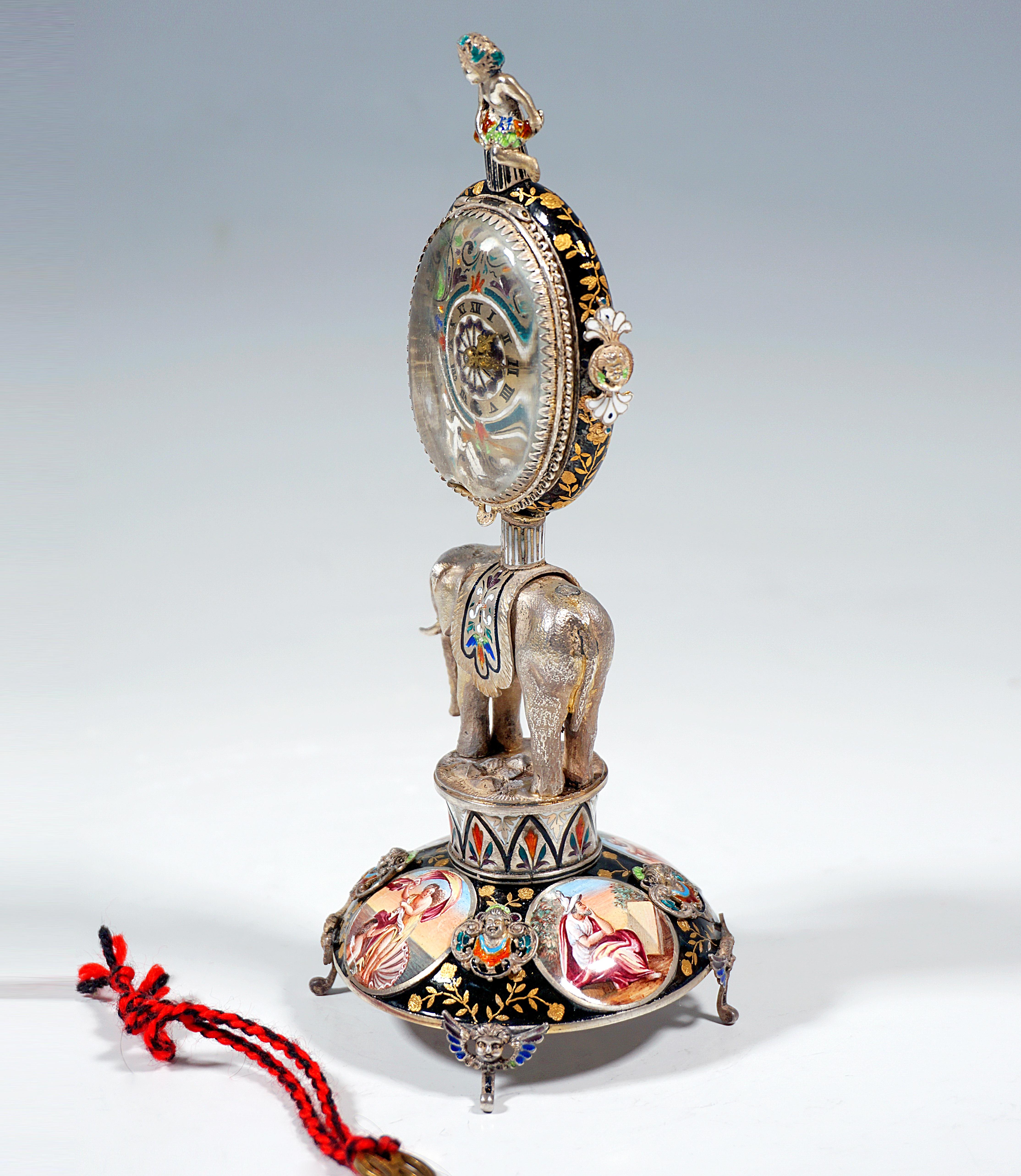 Artfully decorated silver clock by a Viennese master:
Plastic silver elephant on a drum carrying the oval watch case, based on a round, curved base with fine enamel painting - four medallions with antique scenes on a black background with flower