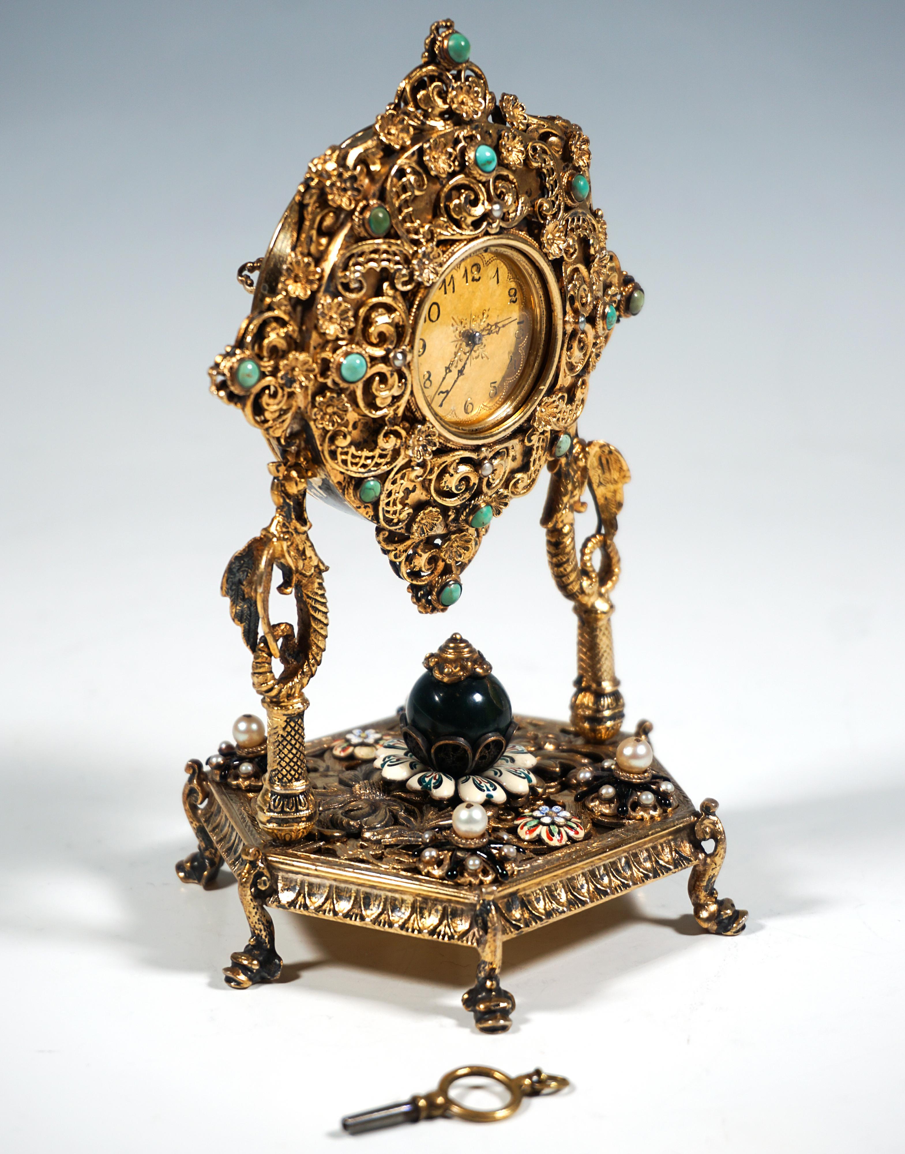 Reloj de sobremesa de plata dorada artísticamente decorado por un maestro vienés:
Placa base plana, en forma de panal, con relieve de palmeta en los lados, que se alza sobre seis pies de delfín adosados a las esquinas, ricamente decorada con