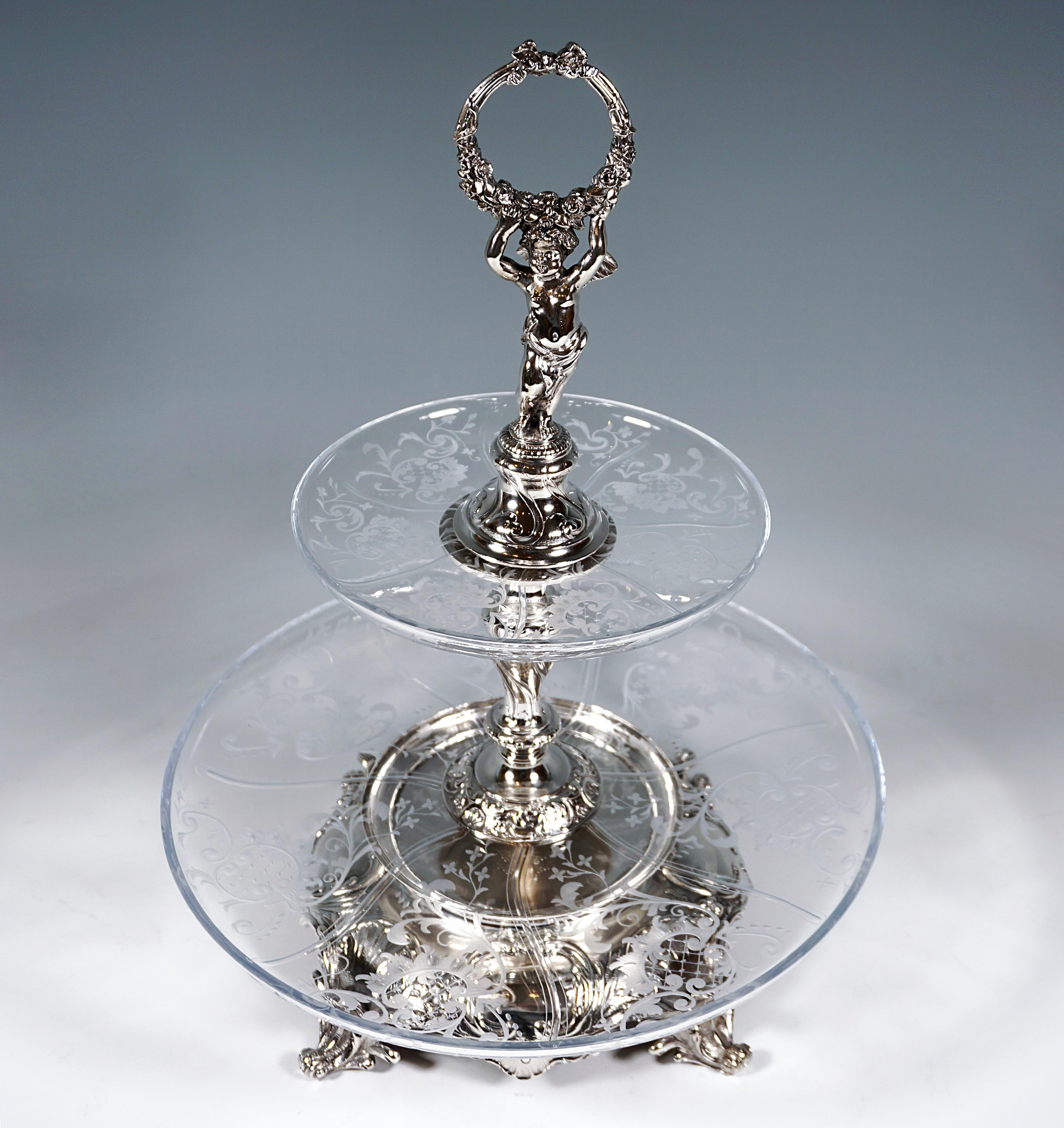 Excellent original Viennese Art Nouveau centerpiece:
Two-tiered, silver table étagère on a large, domed stand richly decorated with volutes and panels on four rocaille feet, baluster-shaped shaft, crowned by a fully sculpted, winged putto on a