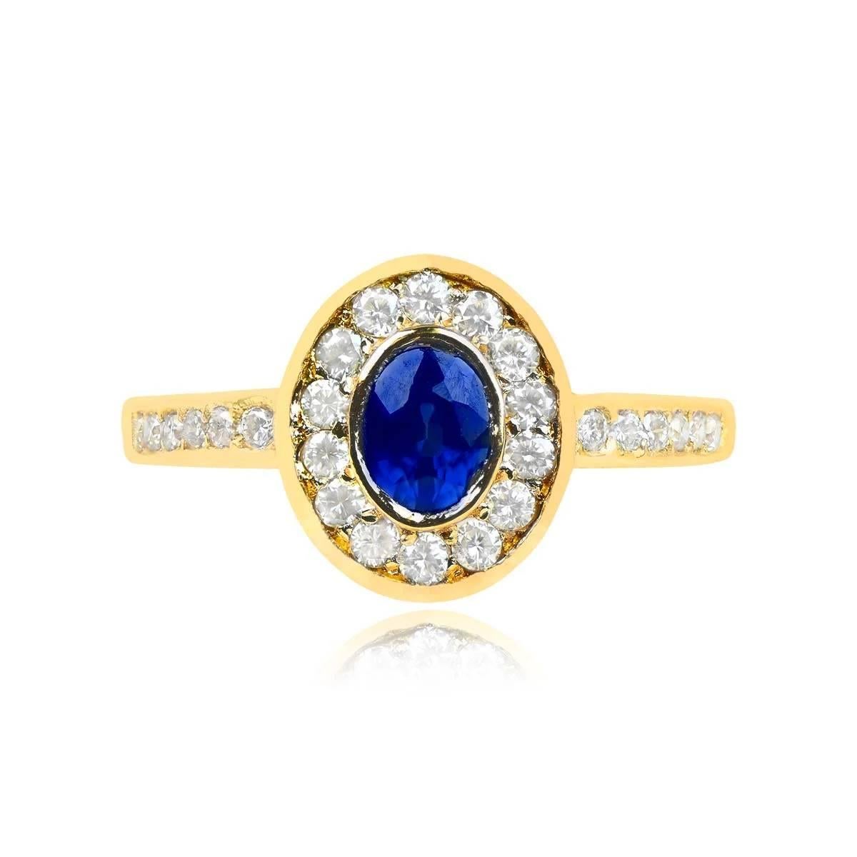 An enchanting vintage French ring showcasing a bezel-set 0.40-carat oval-cut sapphire at the center, surrounded by a halo of round brilliant cut diamonds. The shoulders are adorned with six additional round brilliant-cut diamonds on each side, with