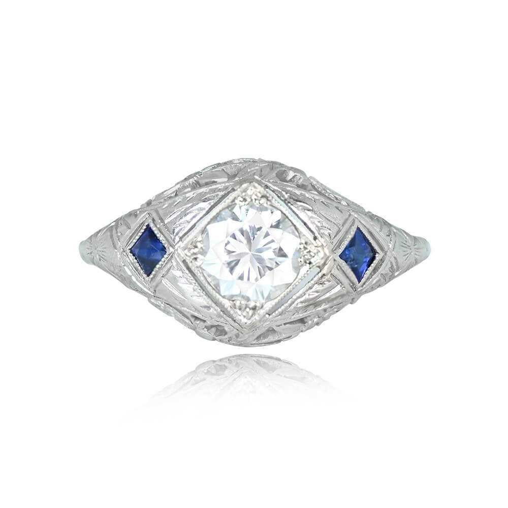 This vintage engagement ring showcases a 0.65-carat round brilliant cut diamond with I color and SI1 clarity. French cut sapphires, totaling about 0.10 carats, adorn each side of the diamond. The ring is adorned with intricate floral and